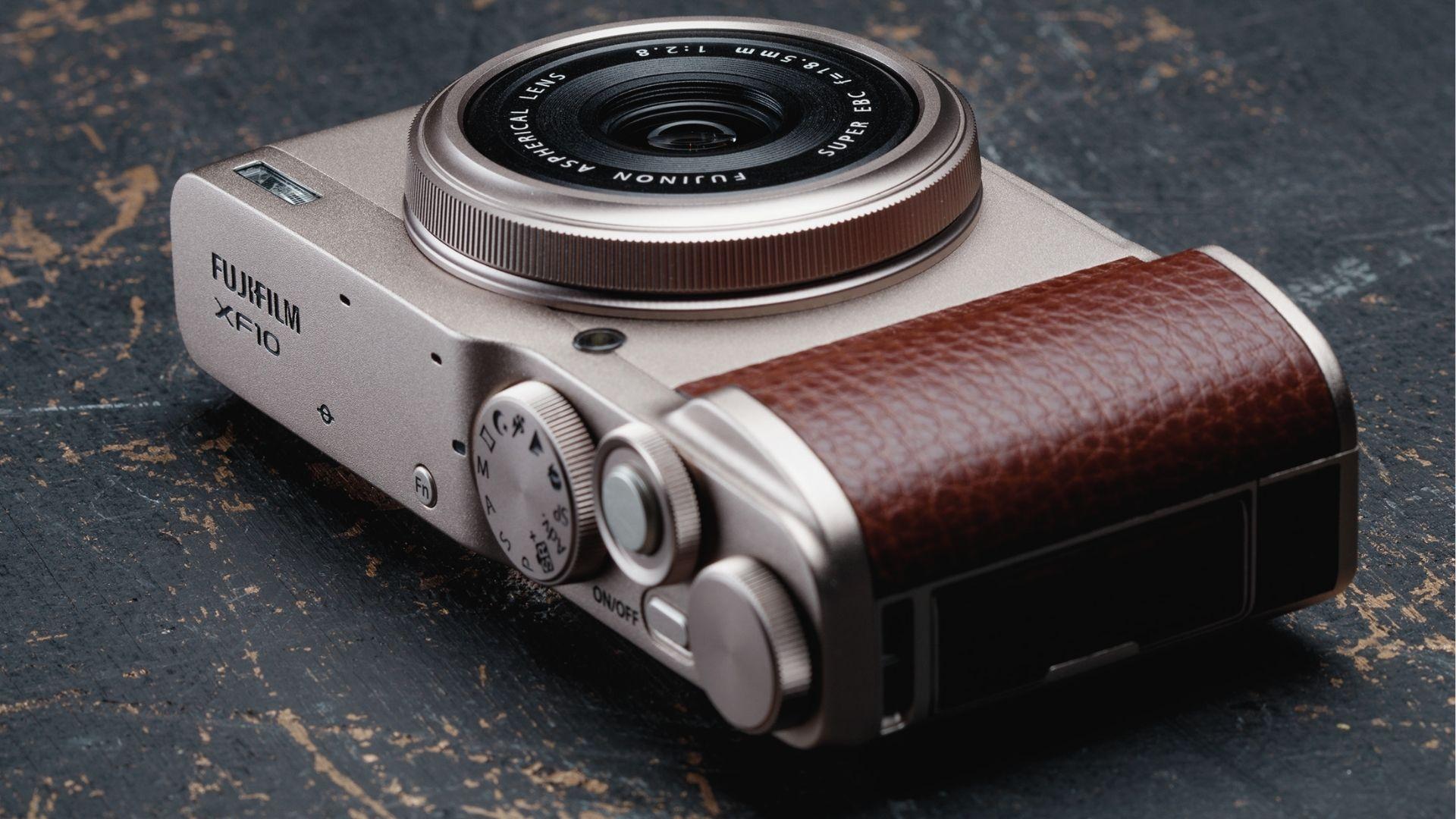 Champagne silver Fujifilm XF10 with brown leather grip on textured dark surface, retro street camera styling.