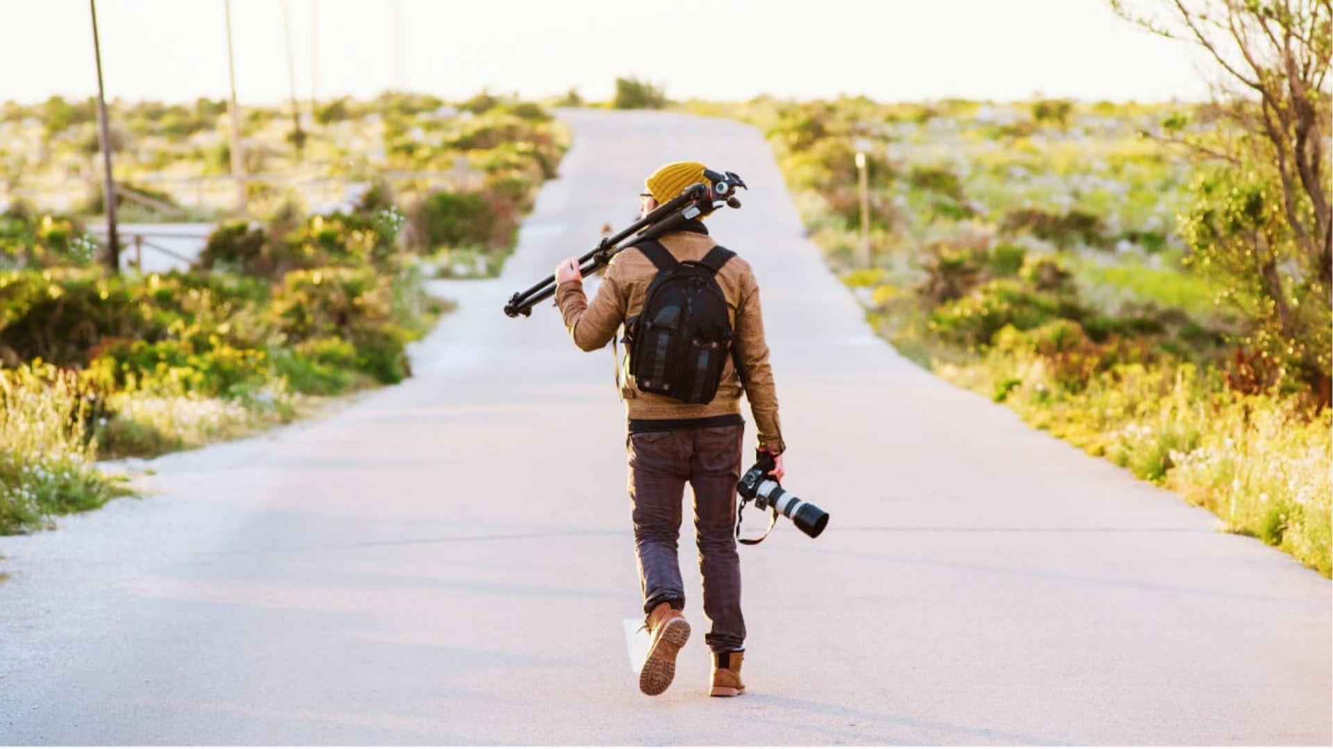 Solo traveler walking down a sunlit country road at golden hour, backpack and tripod on shoulder, DSLR in hand.