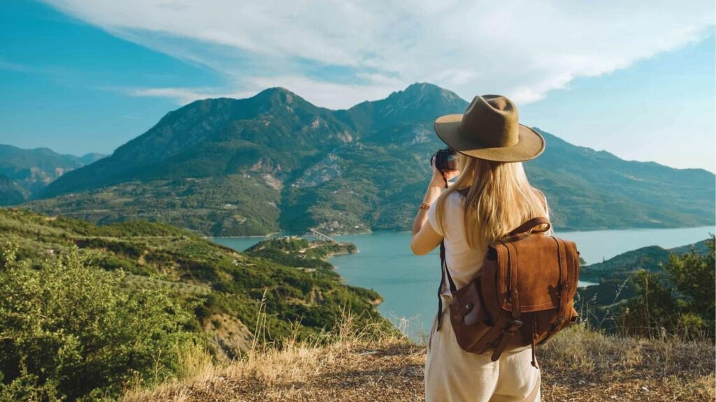 Woman in wide-brim hat and leather backpack photographing a lake and mountain vista with compact camera.