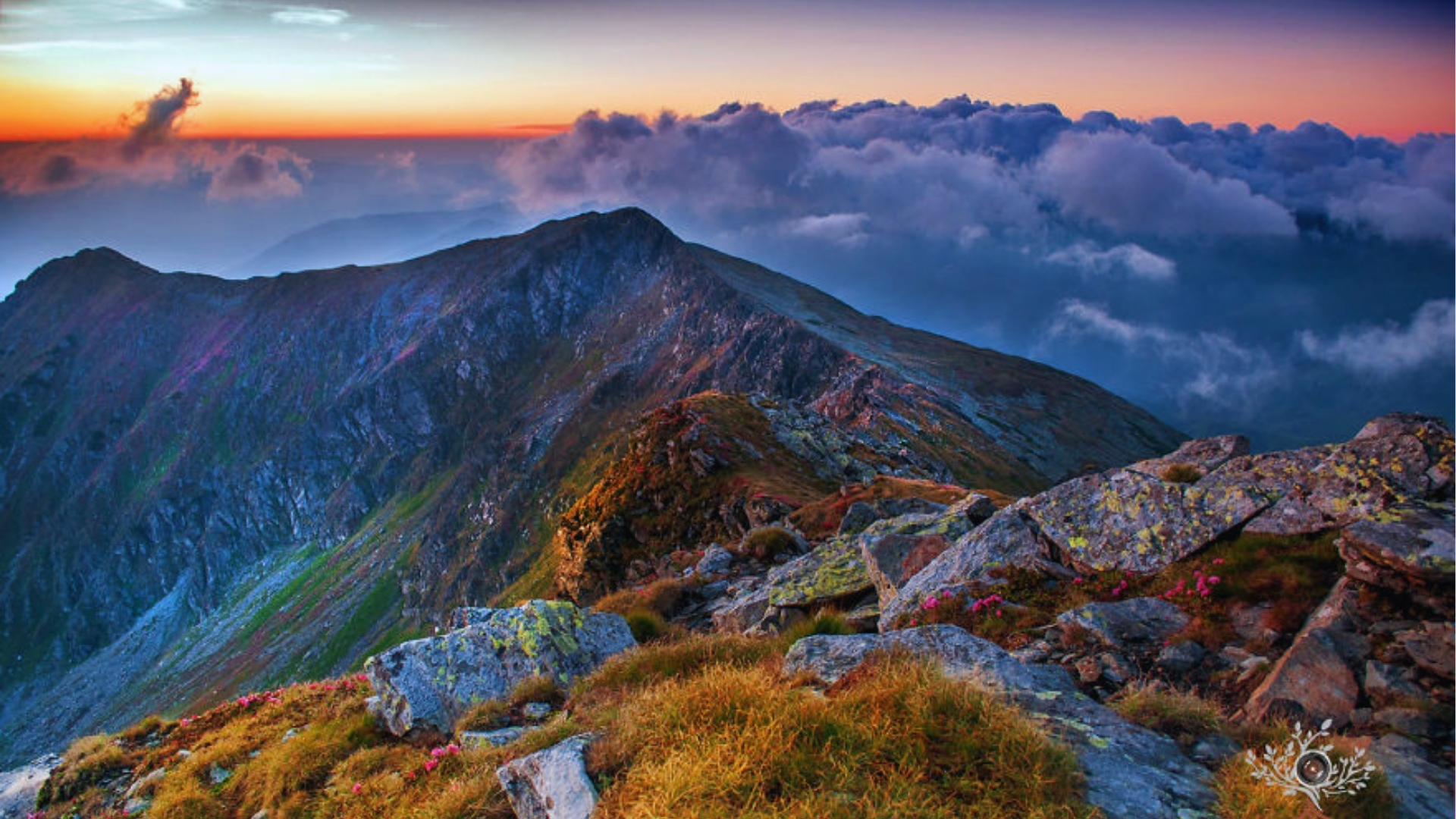 Sunset over a rugged mountain range with rocky peaks and grassy patches in the foreground. Clouds blanket the background, creating a serene, majestic tone.