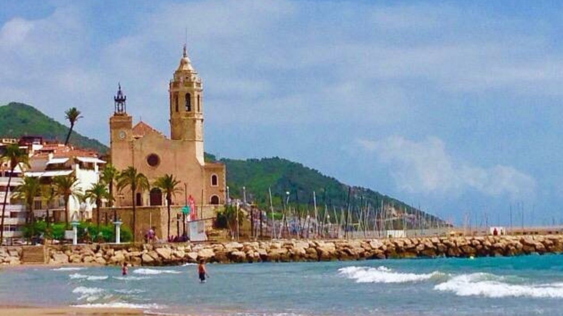 Seaside view of Sitges, Spain, featuring a historic church with a tall bell tower, palm trees, a sandy beach, gentle waves, and moored sailboats in the background.