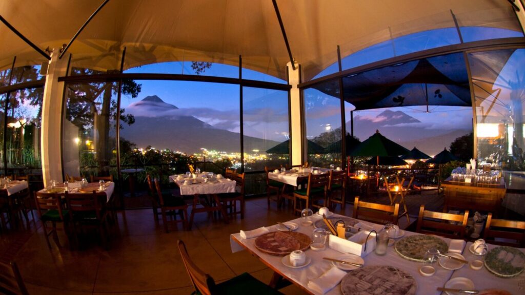 A scenic restaurant interior with panoramic window views of a serene, twilight-lit mountain and cityscape. Tables are elegantly set, suggesting a calm, inviting ambiance.