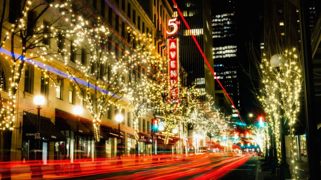 A vibrant city street at night with trees adorned in sparkling white lights, a vivid red "5th Avenue" sign, and colorful light trails from moving cars.