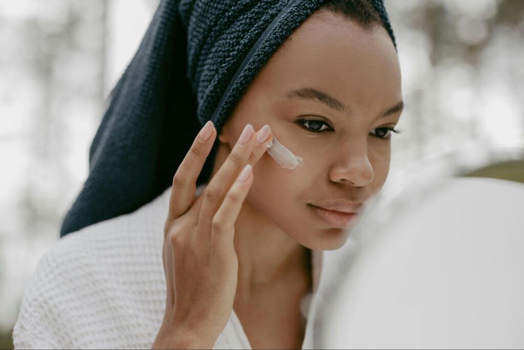 A woman in a towel applies cream to her face, focusing on skincare in a bright, serene bathroom setting.