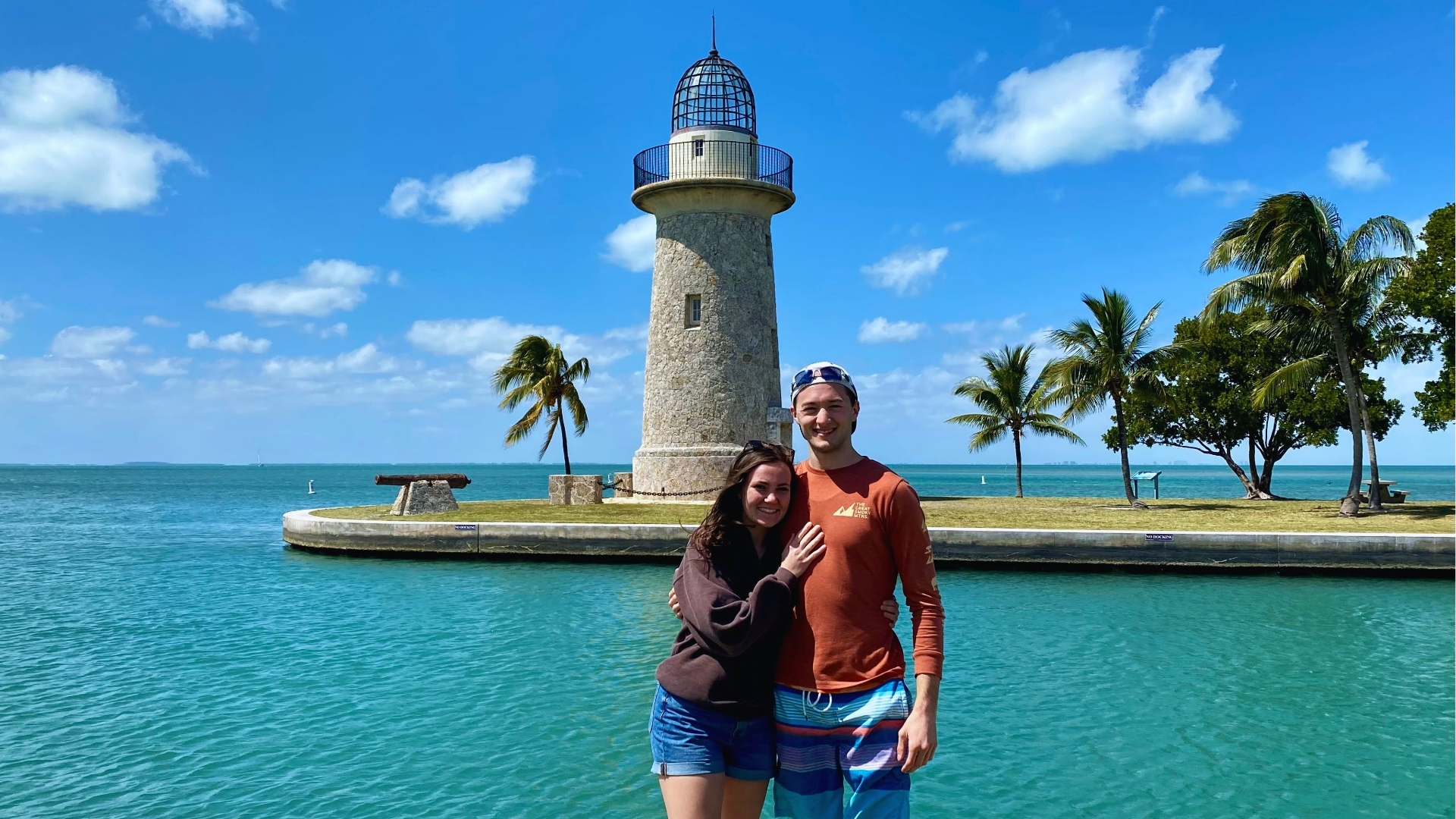 A couple stands smiling in front of a lighthouse by a tropical shoreline. The sky is clear and blue, with palm trees and calm water in the background.