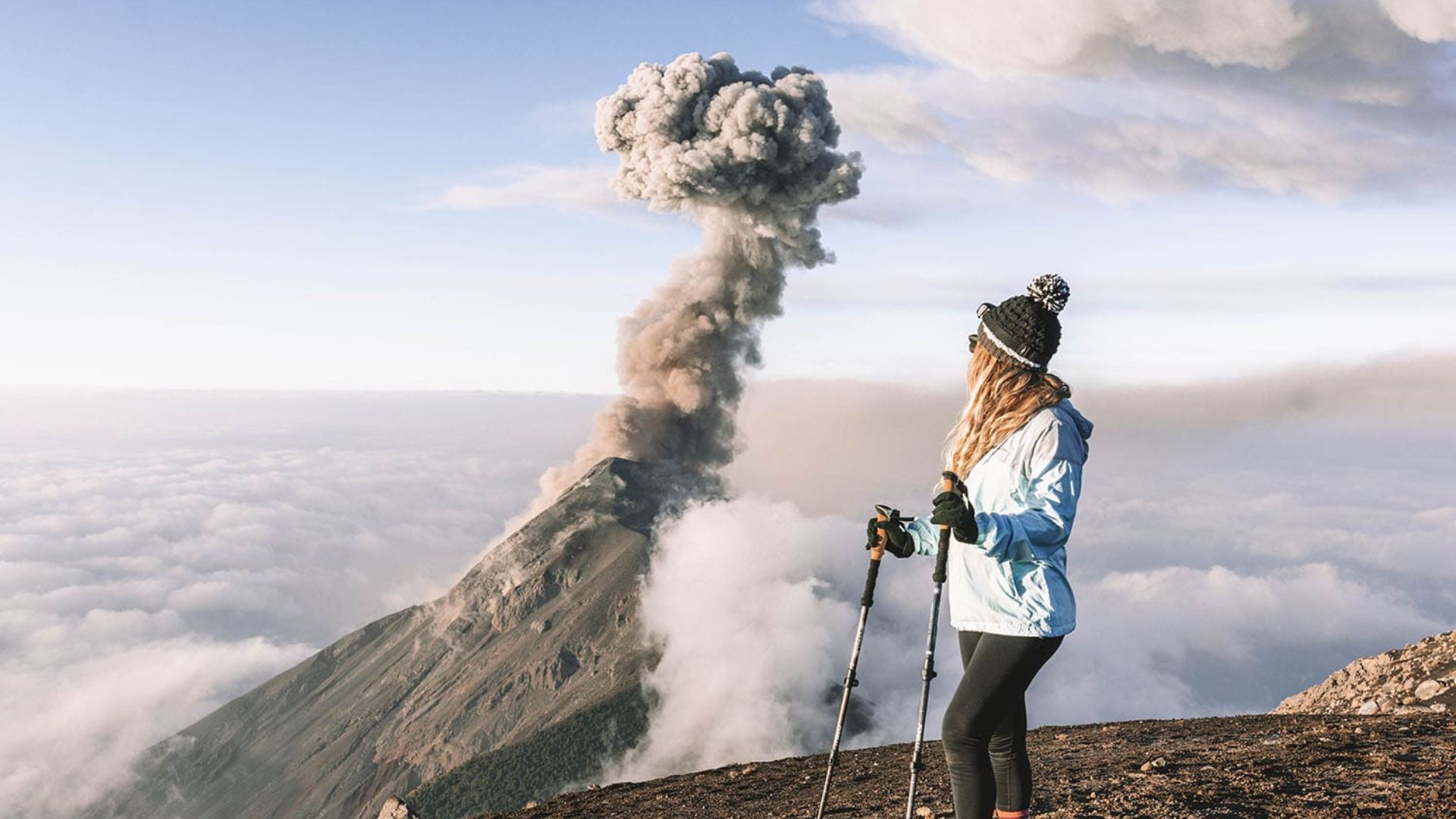 A woman in hiking gear stands on a mountain, gazing at an erupting volcano. Smoke billows into the sky, creating a dramatic and adventurous scene.
