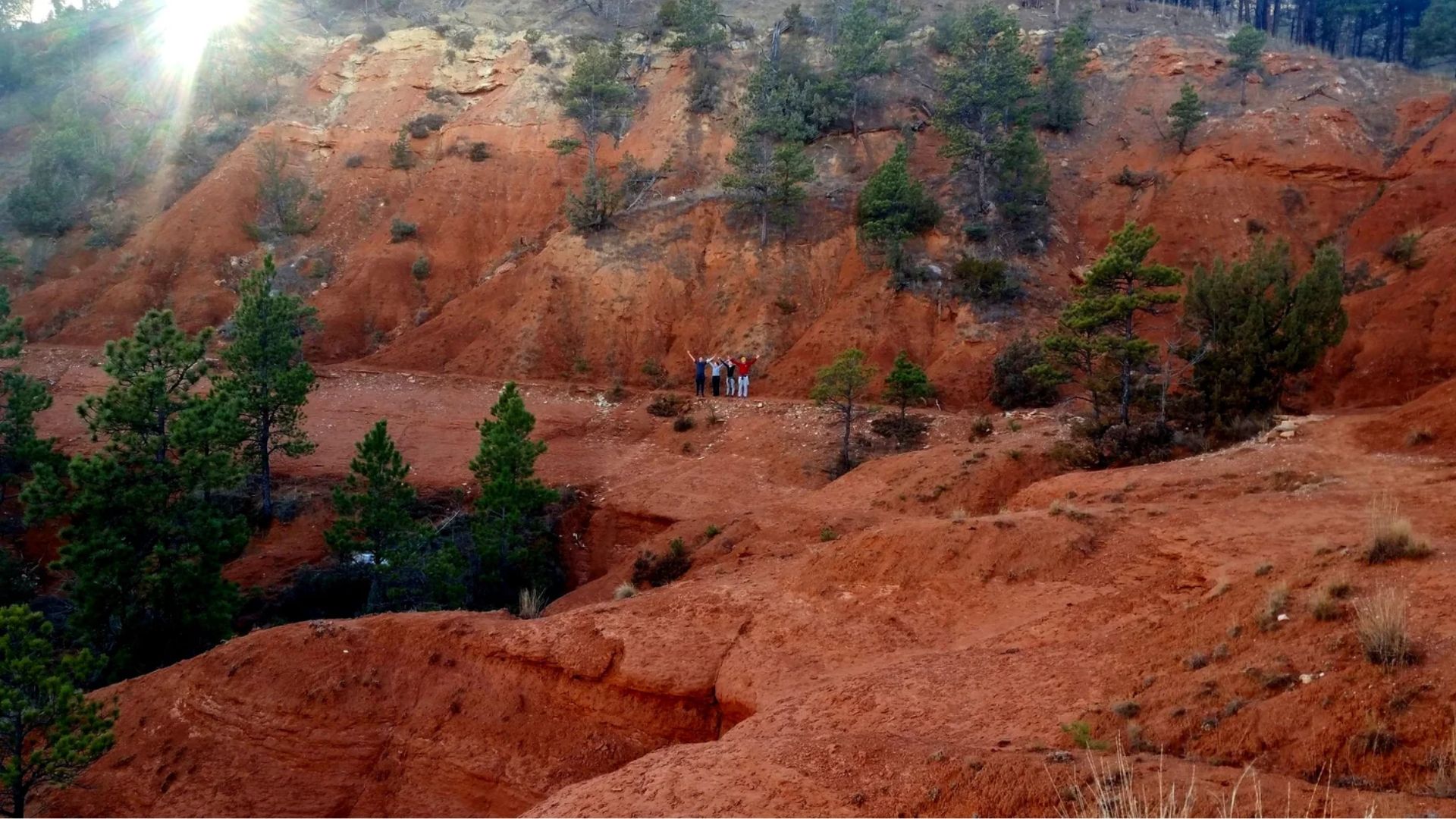 Red rock canyon landscape with scattered pine trees and five people standing on a narrow ledge in the middle distance. Sunlight filters through the top left.