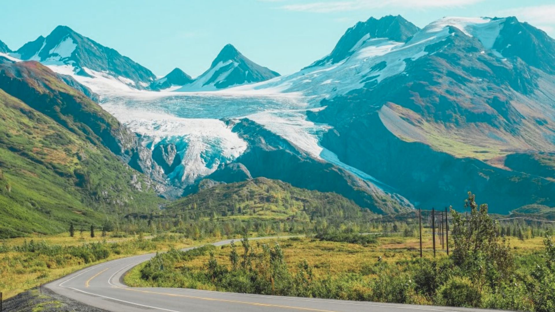 A winding road leads towards majestic snow-capped mountains under a clear blue sky. Lush greenery surrounds the scene, evoking a sense of peace.