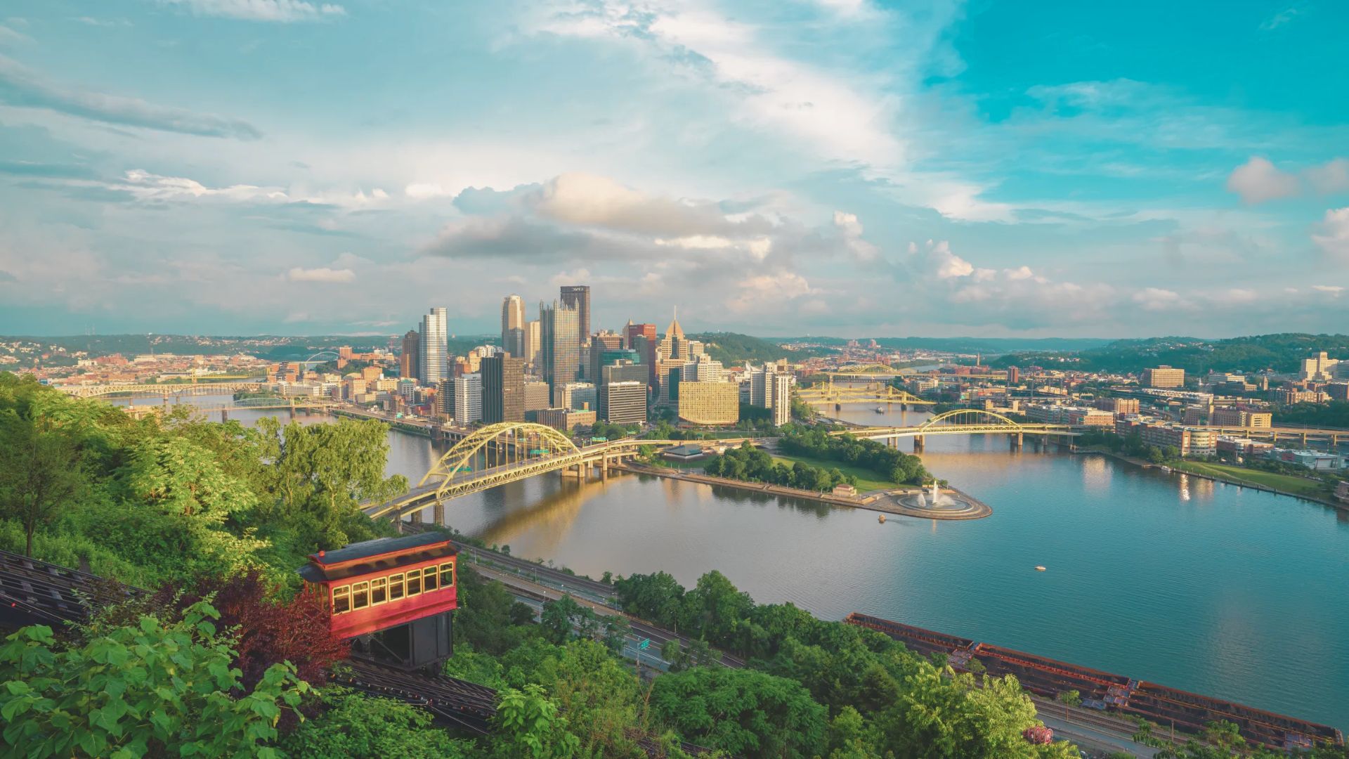 Scenic view of a city skyline surrounded by rivers and bridges under a bright blue sky. A red cable car travels up a lush, green hillside.