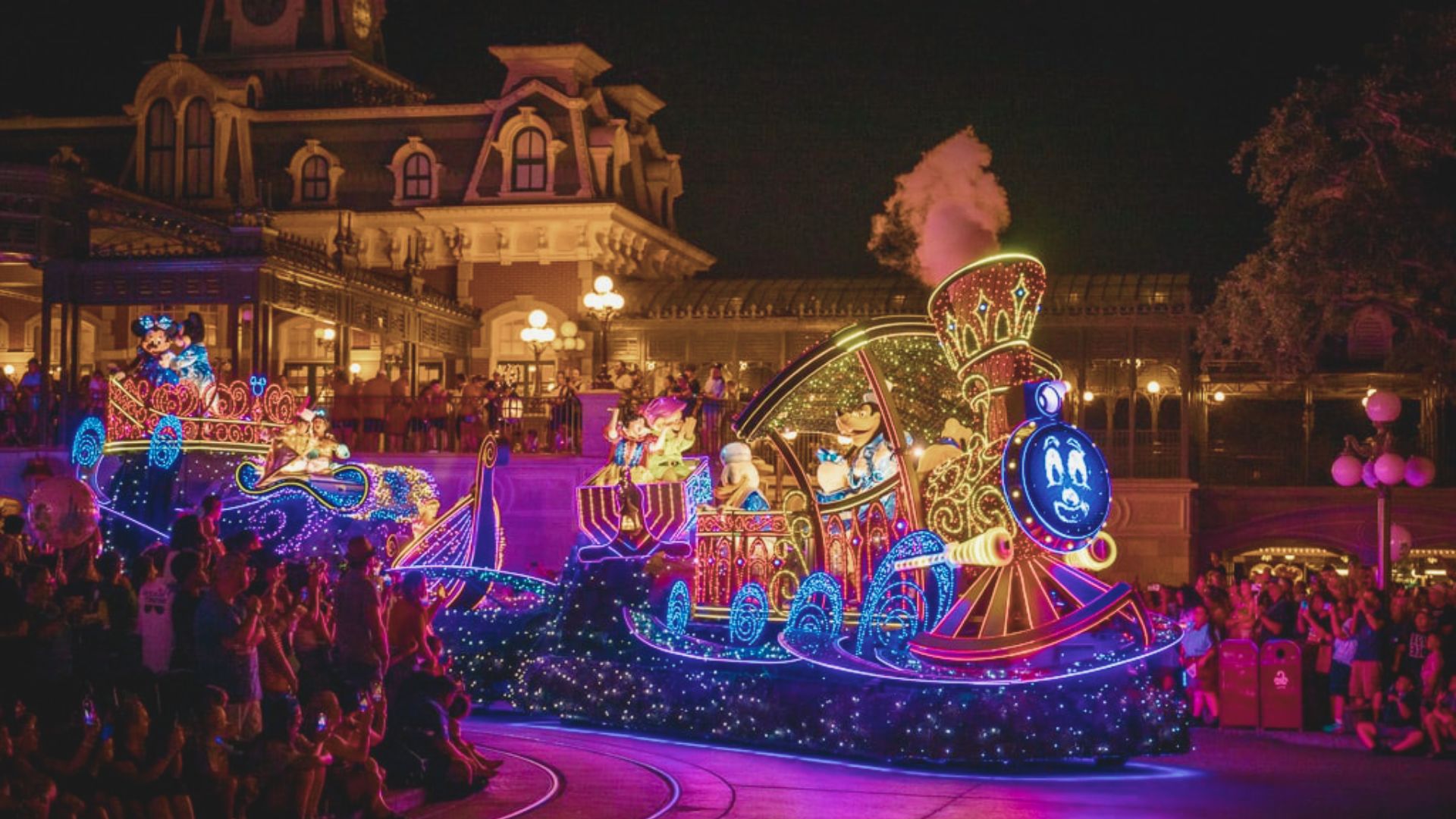A vibrant nighttime parade features an illuminated train with characters, colorful lights, and a cheering crowd, conveying joy and excitement in a festive setting.