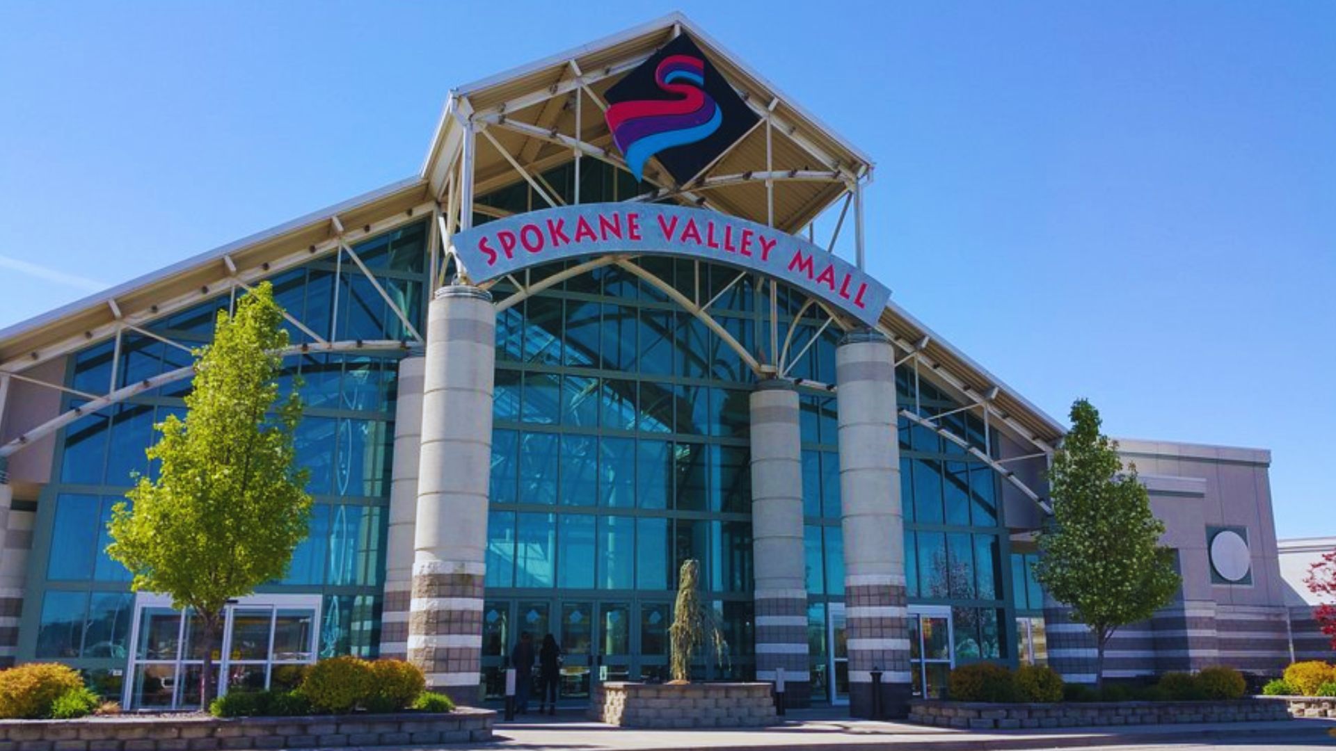 Front entrance of a large shopping center featuring glass doors and a wide walkway, bustling with shoppers.