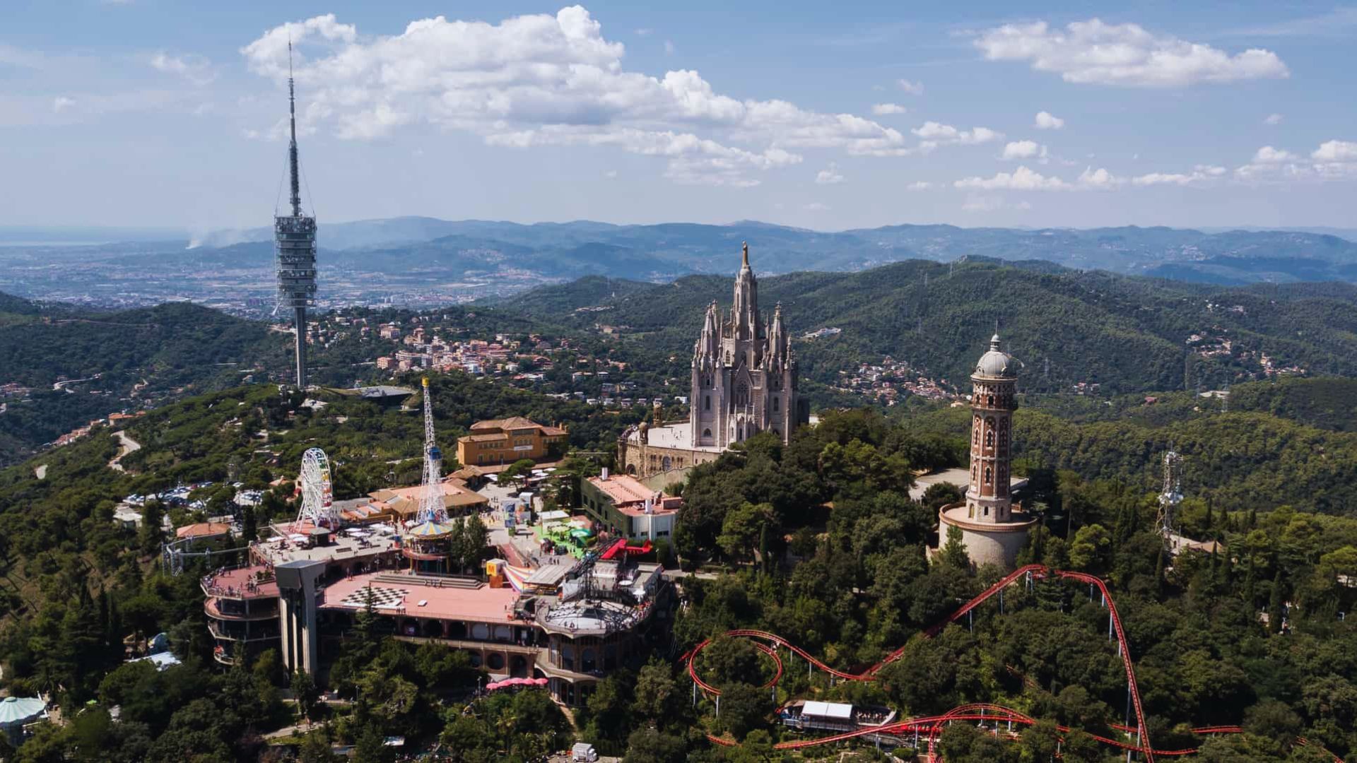 Aerial view of Tibidabo Amusement Park with a roller coaster, surrounded by lush greenery. In the background, the Temple Expiatori del Sagrat Cor and a tall communications tower are visible against a backdrop of distant mountains and a partly cloudy sky.