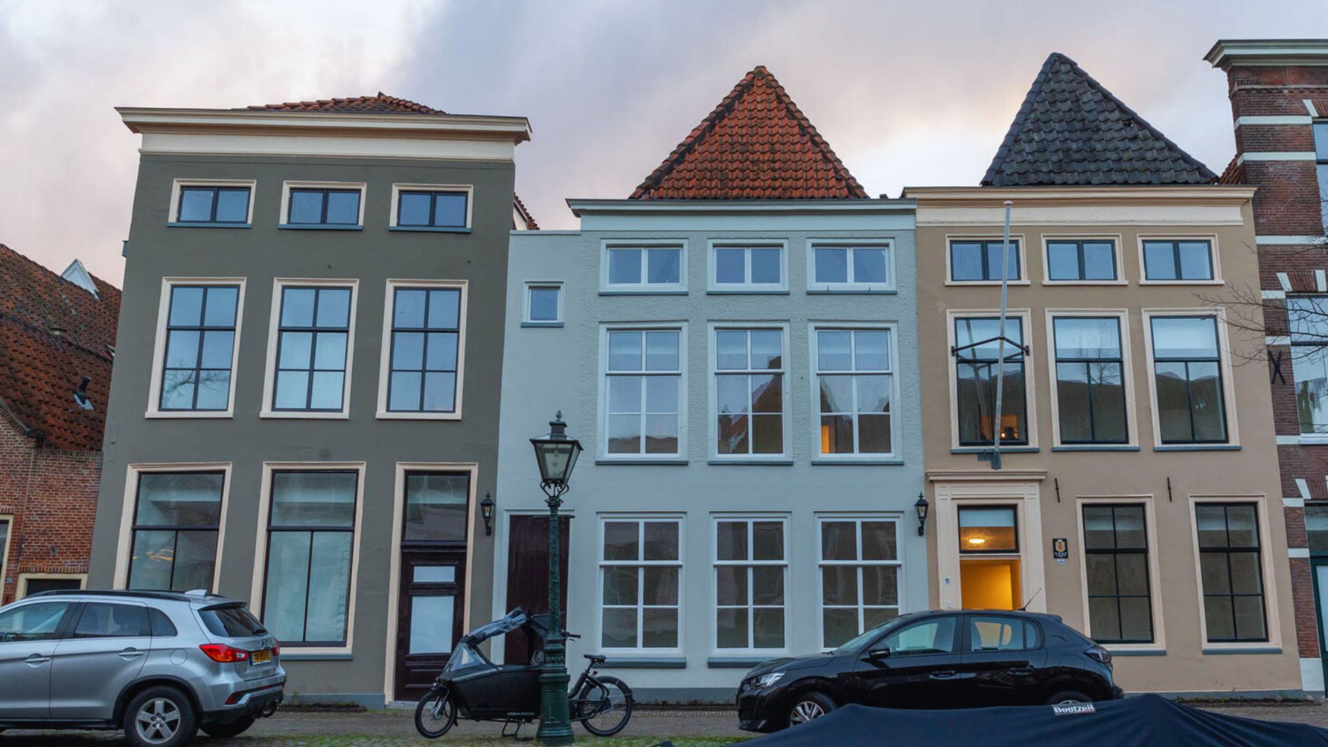 A row of three narrow, tall Dutch-style houses with large rectangular windows and triangular roofs. Cars and a bicycle are parked in front.