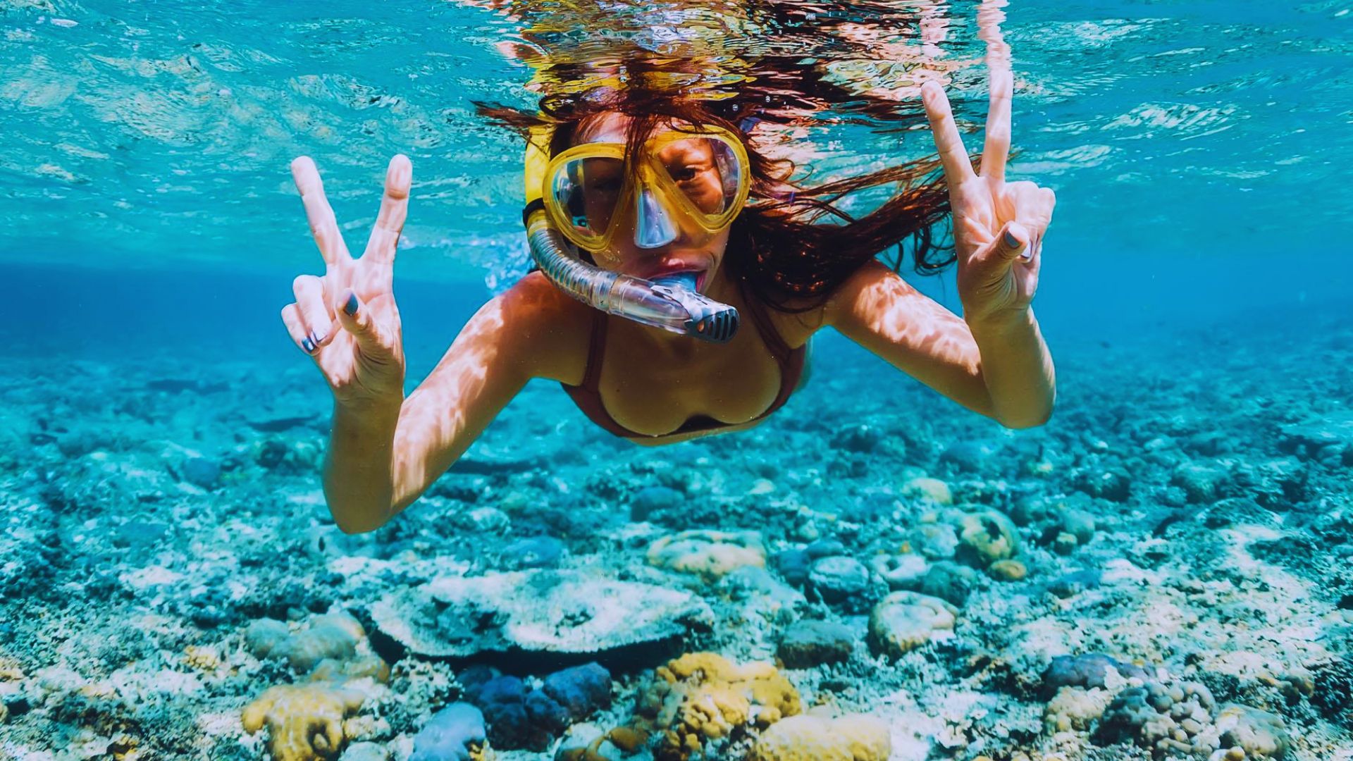 Underwater view of a woman snorkeling over a vibrant coral reef. She wears goggles, a snorkel, and shows peace signs with both hands, exuding joy and adventure.