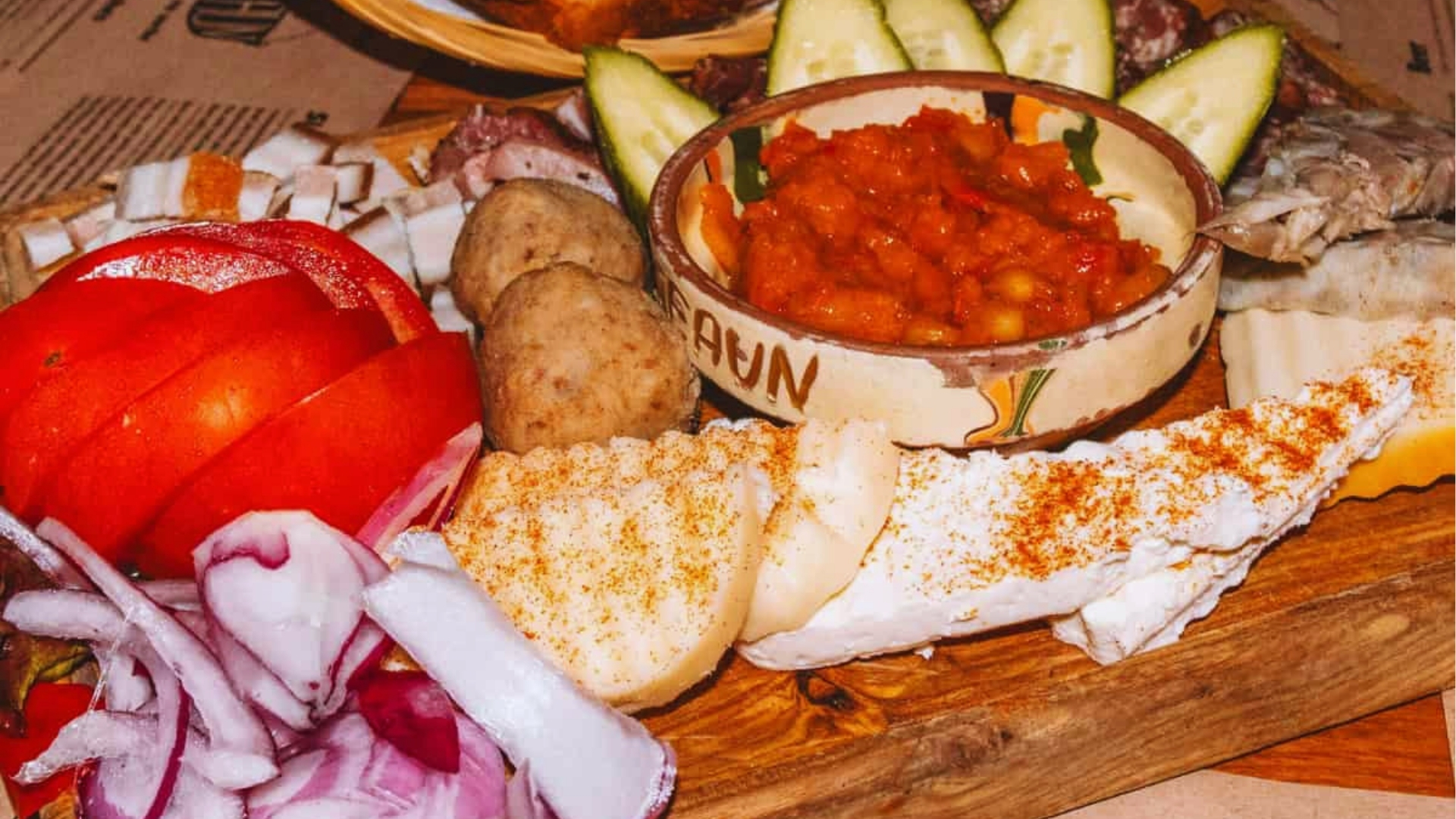 A rustic wooden platter features sliced tomatoes, red onions, boiled potatoes, cucumbers, and a spiced cheese wedge. Central bowl contains red sauce. Cozy, hearty meal.