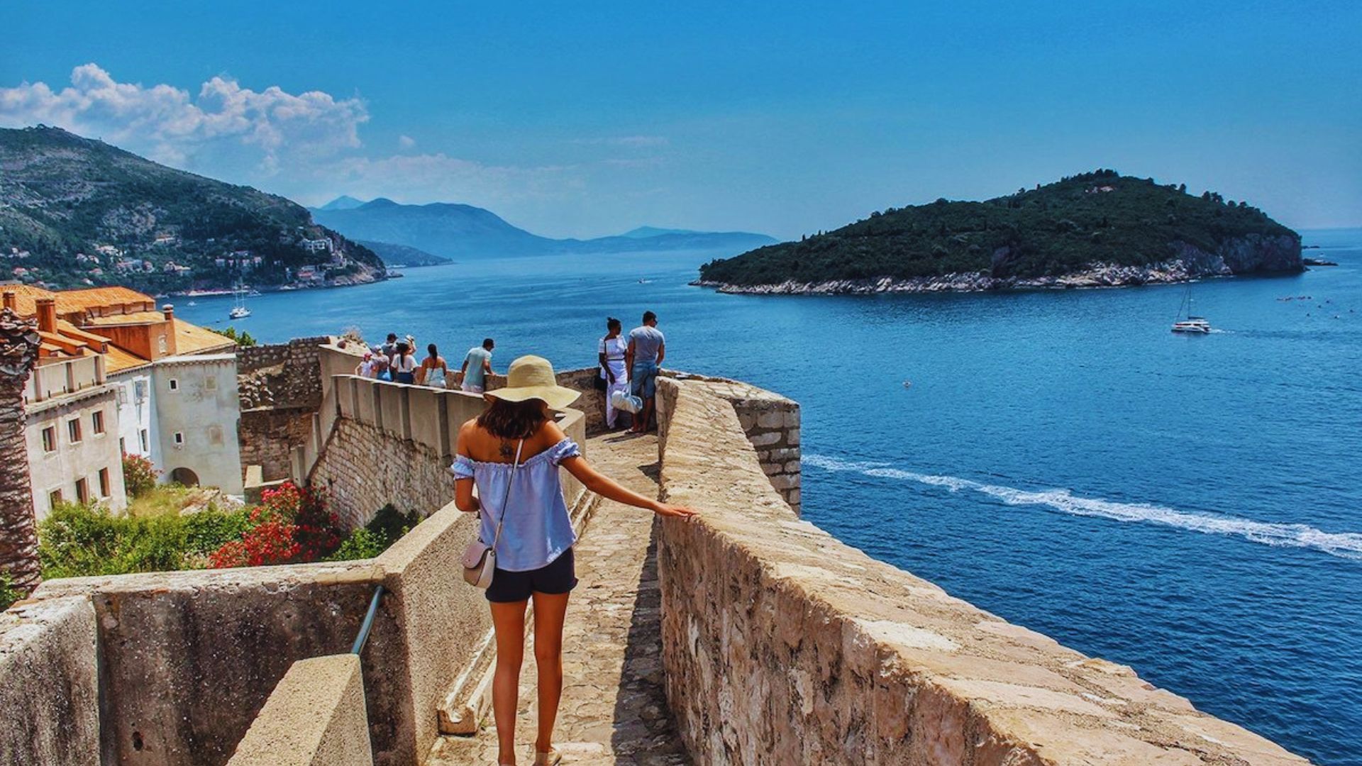 A woman in a sunhat walks along a stone wall overlooking the blue sea and lush islands. Tourists enjoy the sunny day, creating a vibrant, scenic mood.