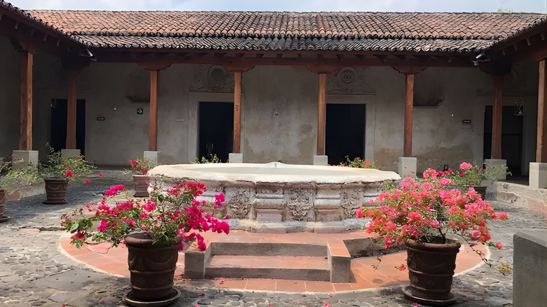 A serene courtyard features a central stone fountain surrounded by vibrant pink flowers in pots. Rustic wooden beams, clay tiles, and muted walls create a historic ambiance.