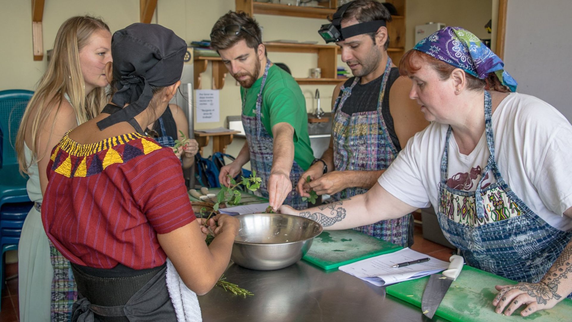 Five people in a kitchen, wearing aprons, attentively prepare ingredients around a table. The atmosphere is collaborative and focused.