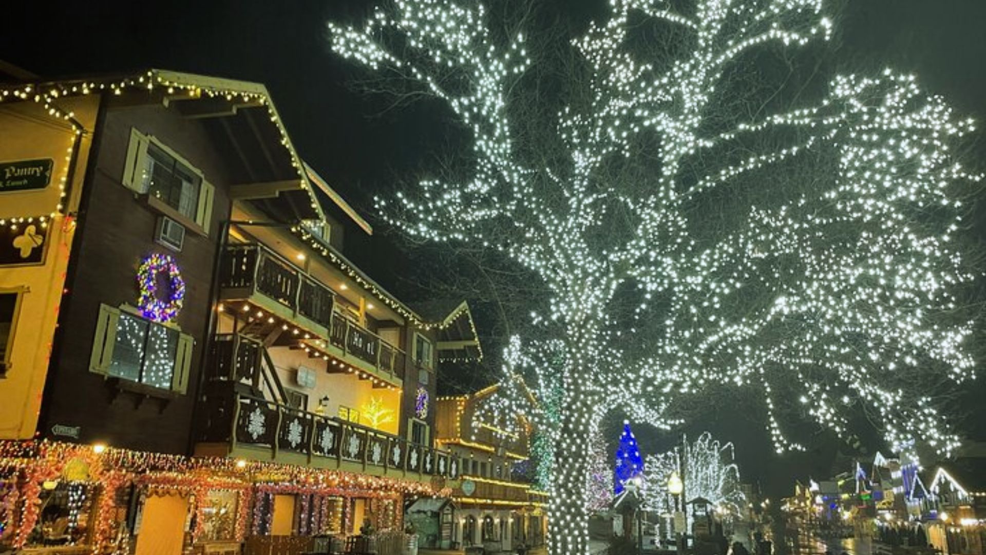 Festive village street at night with buildings adorned in colorful string lights, a large tree sparkling with white lights, evoking a joyful, holiday atmosphere.