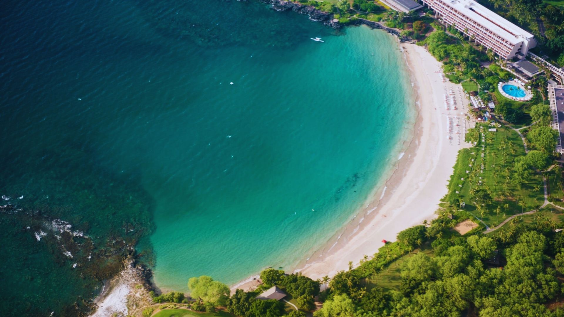 Aerial view of a beach with golden sand and a resort, showcasing clear blue water and lush greenery surrounding the area.