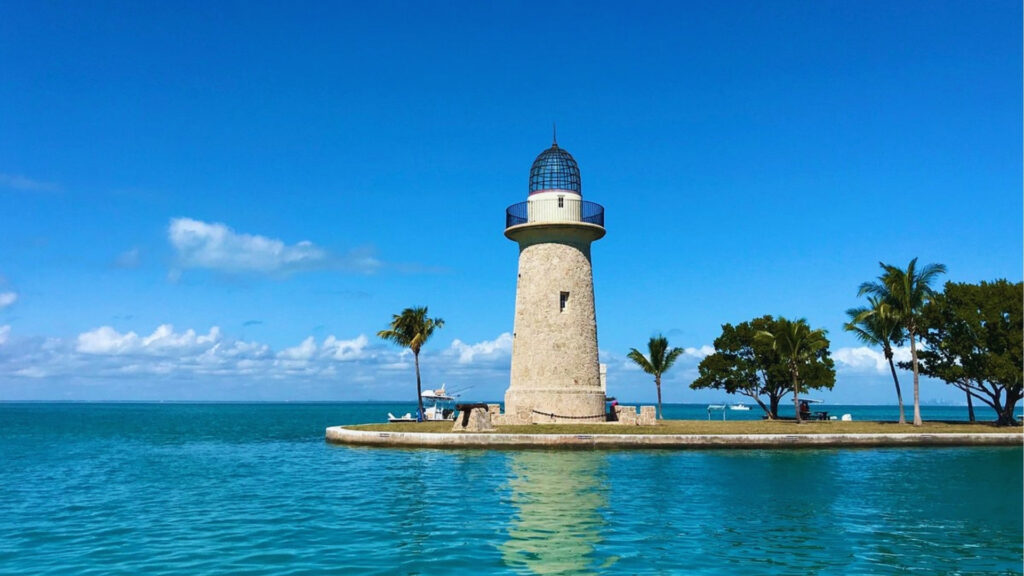A stone lighthouse with a metal dome stands on a small islet surrounded by turquoise water. Palm trees and a clear blue sky create a serene scene.