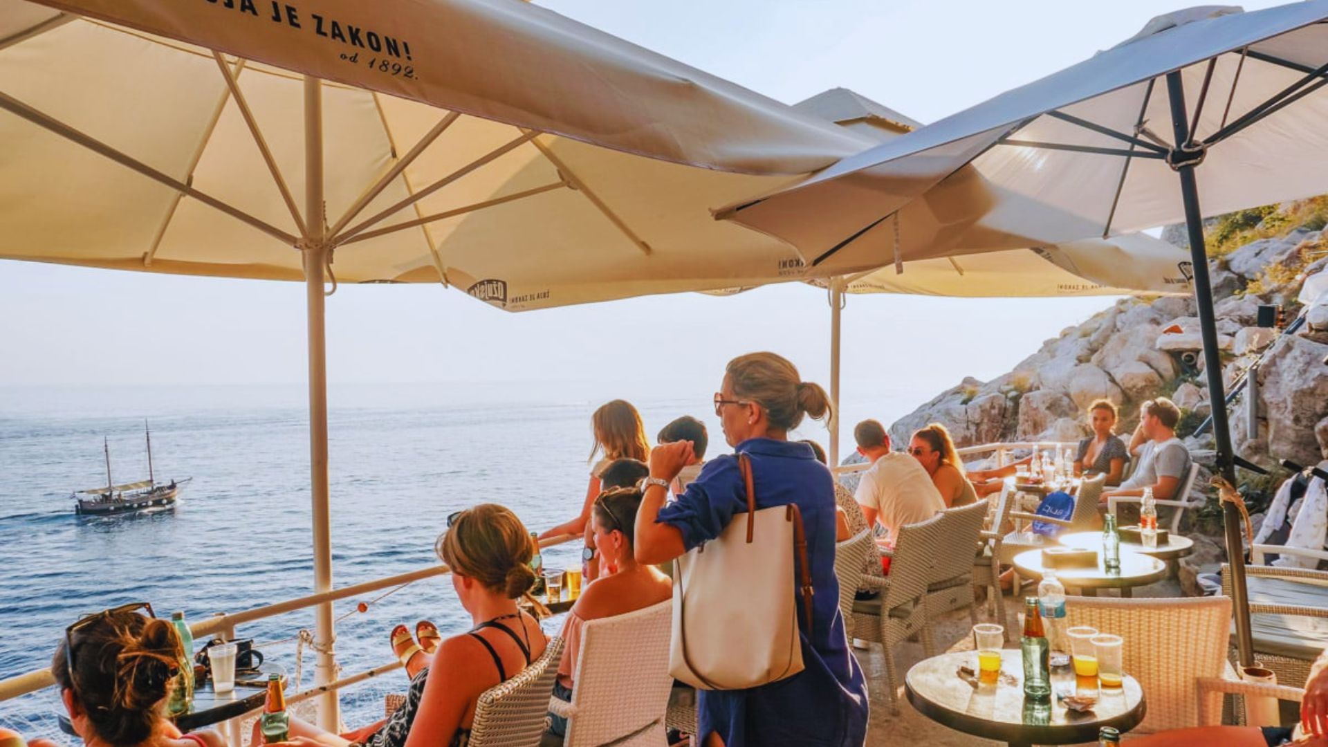 People relaxing under large umbrellas at a scenic seaside café overlooking the ocean. A sailboat is visible in the distance, creating a tranquil atmosphere.