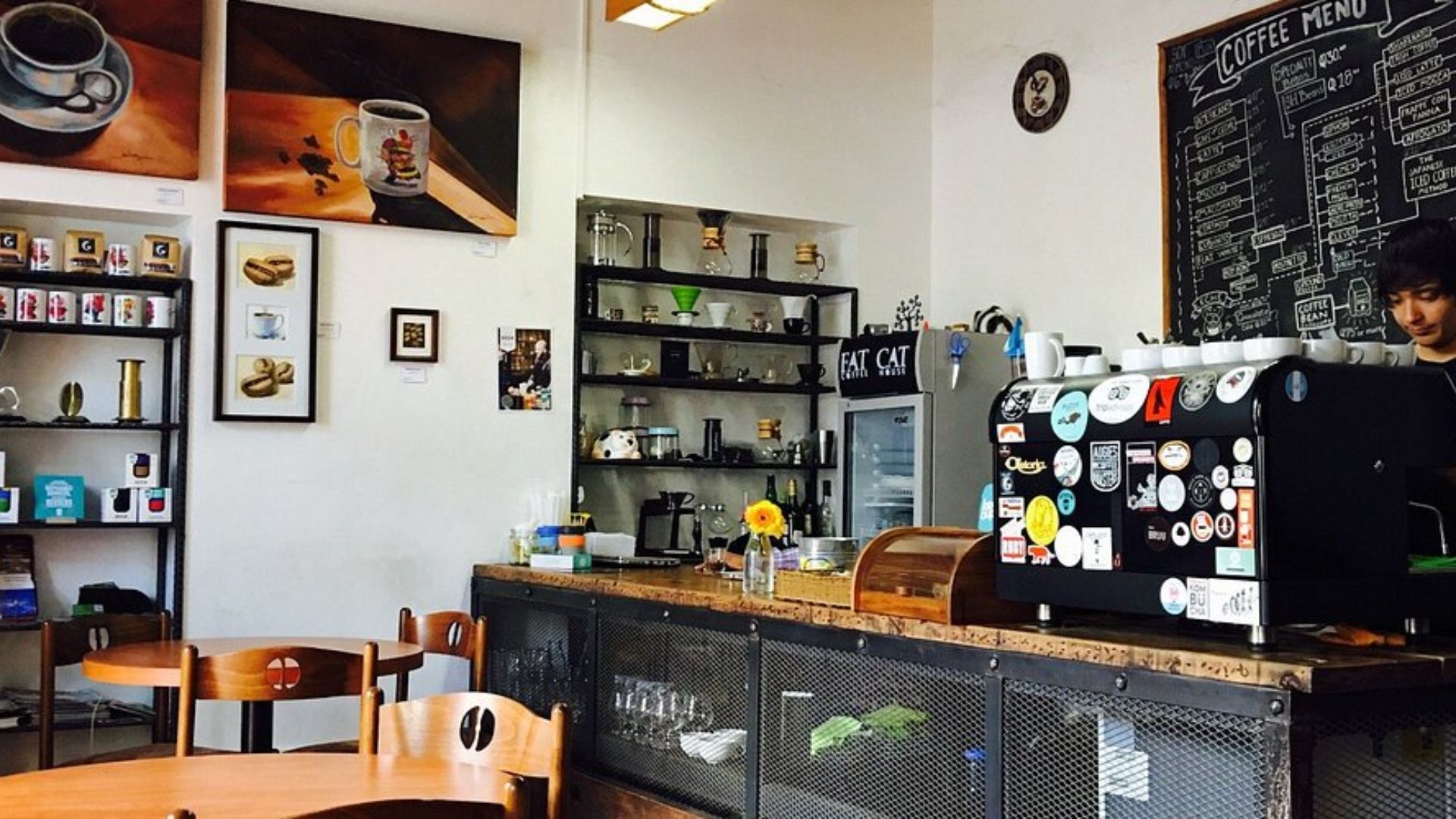 Cozy café interior with a wooden counter adorned with sticker-covered coffee machines. Chalkboard menu, shelves with mugs, and a sunflower in a vase. Relaxed ambiance.