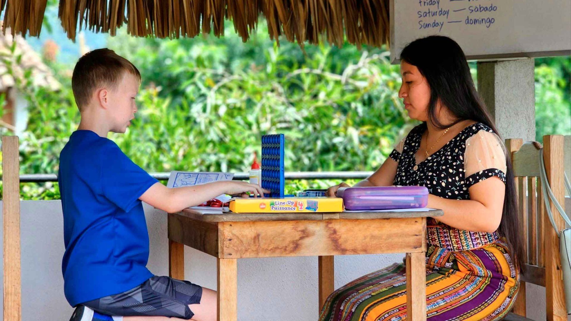 A young boy and a woman sit at a wooden table outdoors, playing a board game. They appear focused, surrounded by lush greenery and a thatched roof.