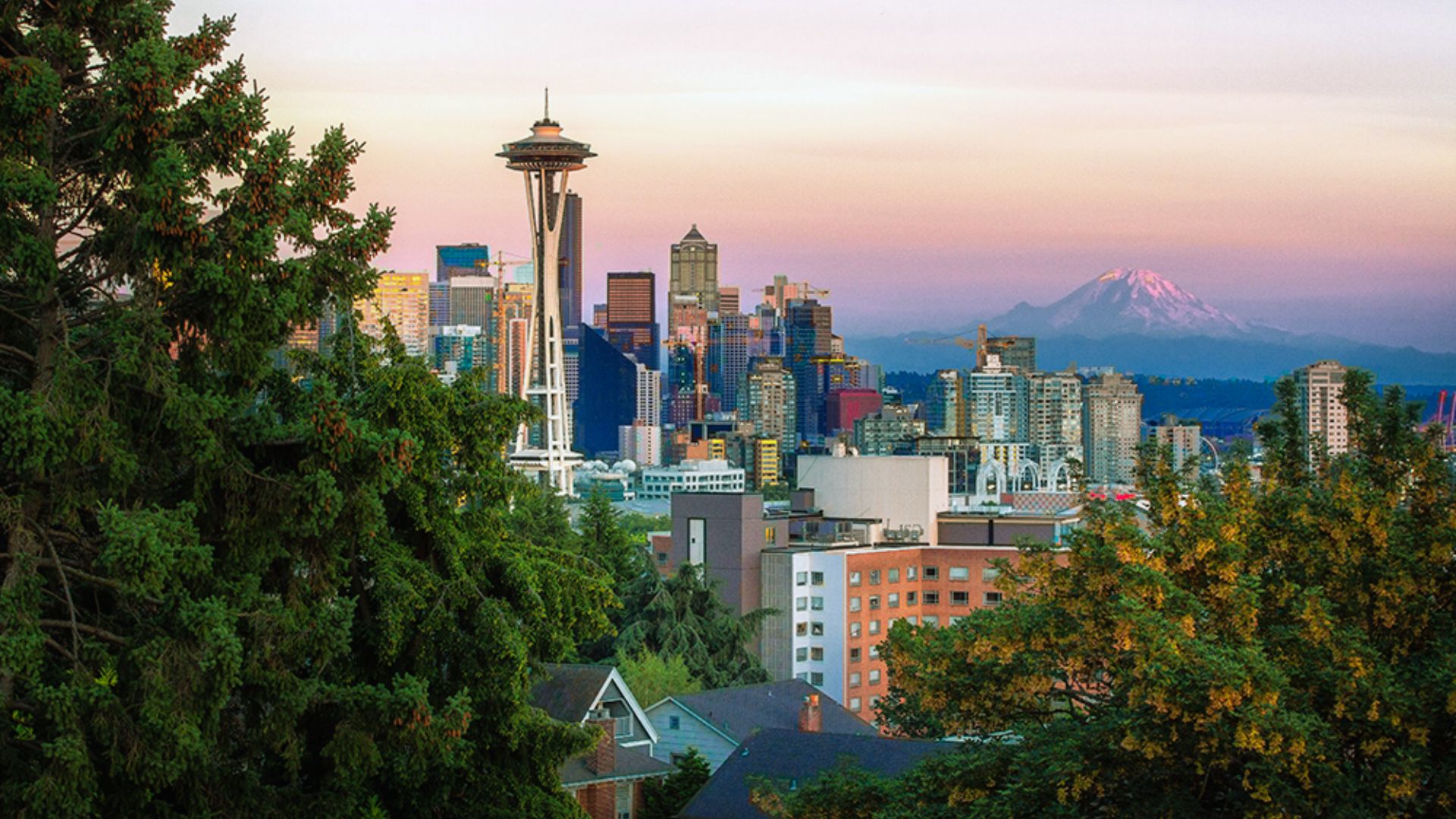Seattle skyline at sunset with the Space Needle prominent amid skyscrapers. Mount Rainier is visible in the background, framed by lush greenery.