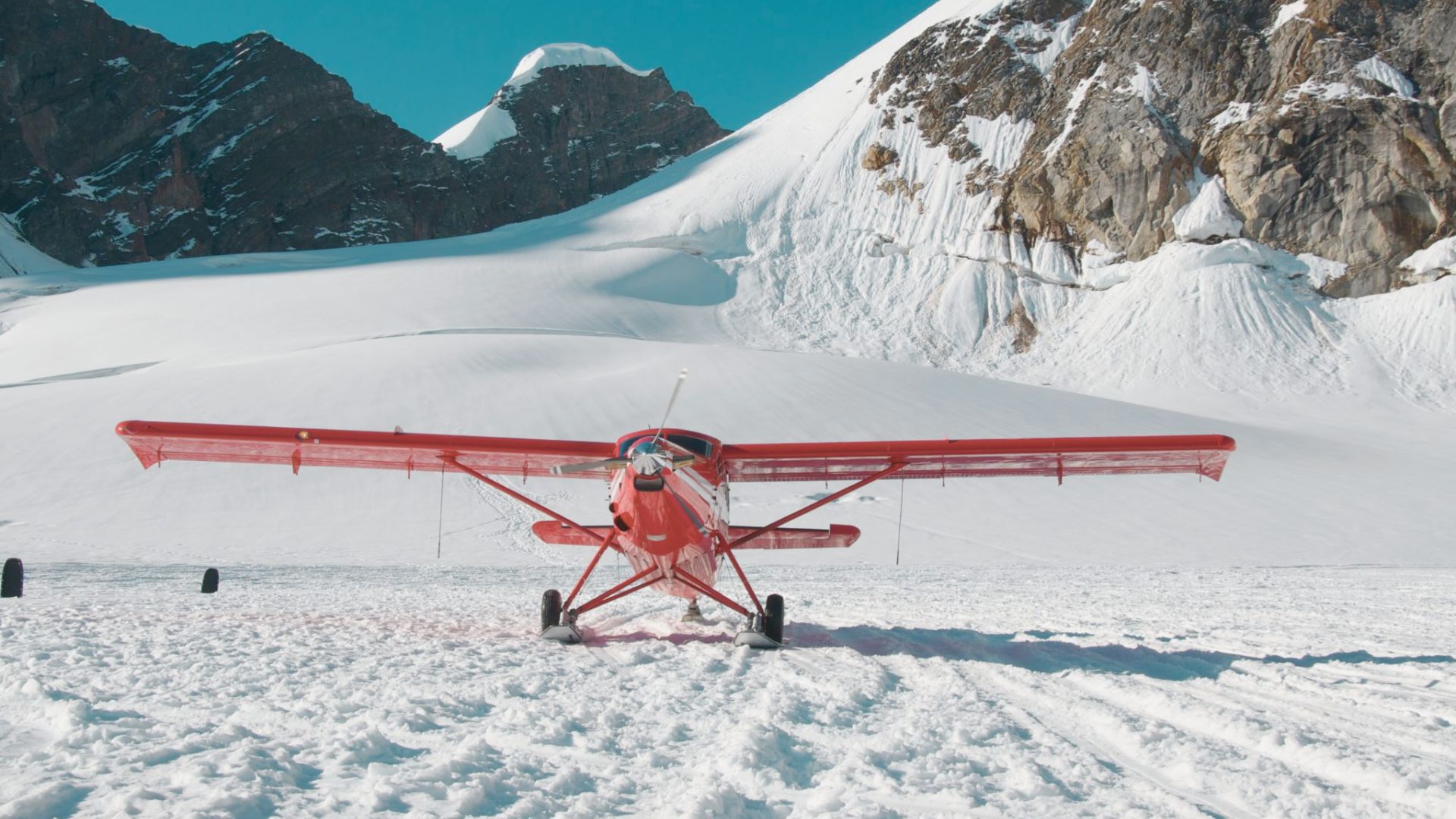 A red small aircraft is parked on a snowy mountain landscape with clear blue sky, surrounded by rocky peaks and snow, conveying a sense of adventure.