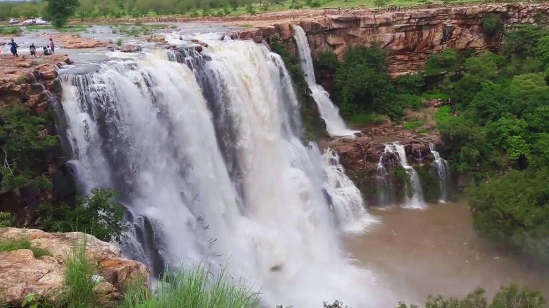 A large waterfall cascades down, with several people standing at the edge, admiring the view.