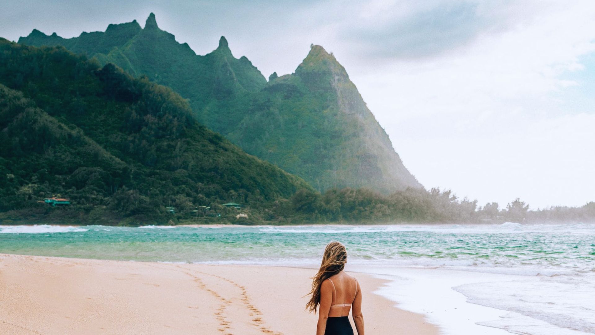 A woman in a bikini stands on the beach, with mountains rising in the background under a clear blue sky.
