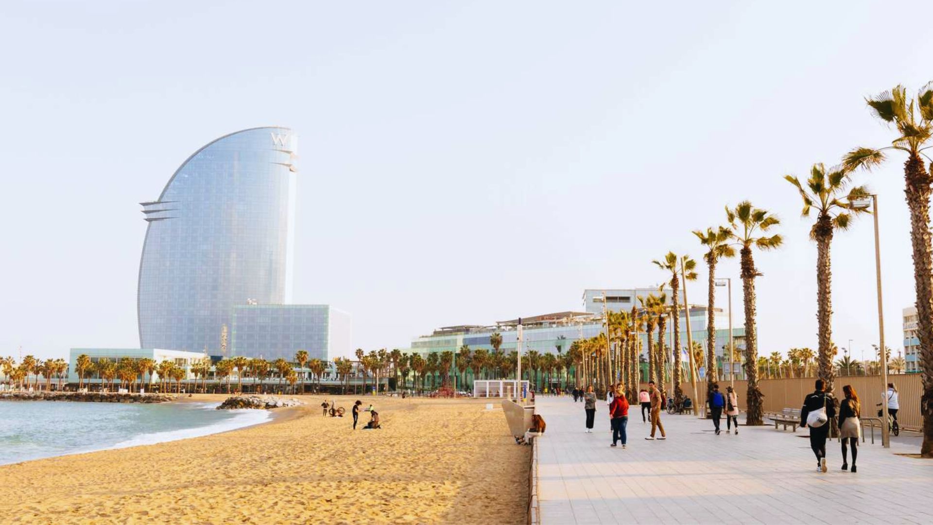 Barcelona beach scene with golden sand, palm trees, and a modern sail-shaped hotel. People stroll along the promenade under a clear blue sky.