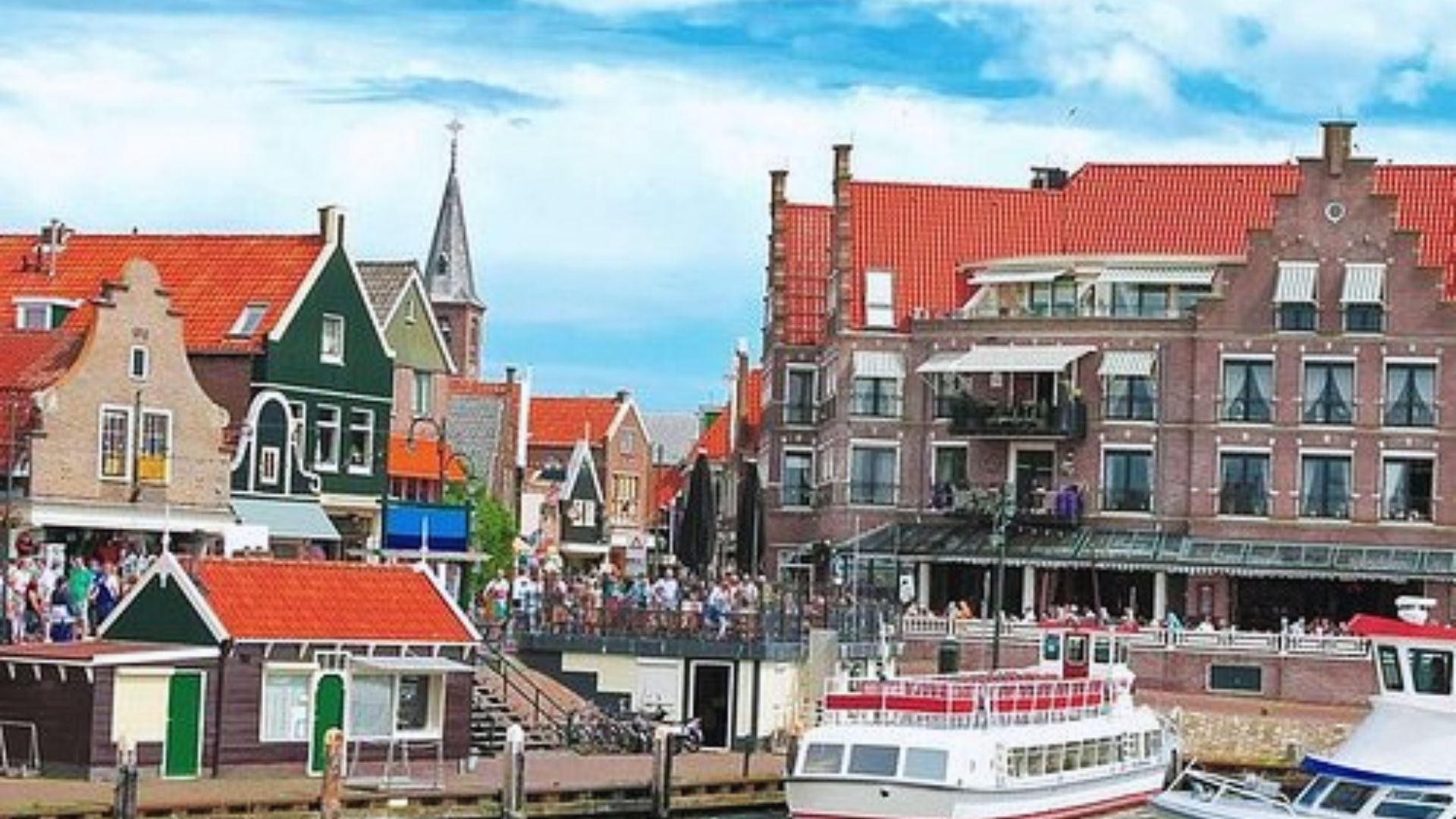 A picturesque harbor scene with colorful Dutch-style buildings featuring steep gables and red roofs. Boats are docked nearby under a bright, blue sky.