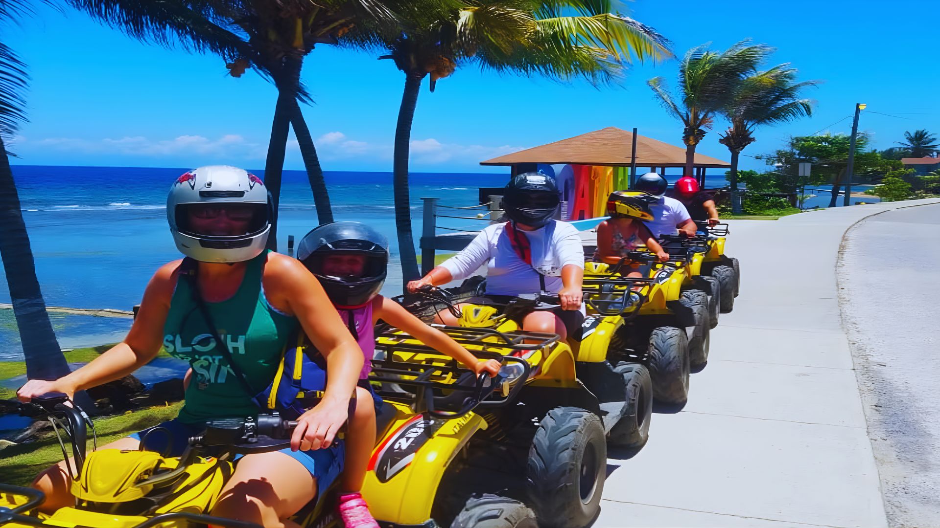 A group of people rides yellow ATVs along a coastal path lined with palm trees, with the ocean and a bright blue sky in the background.