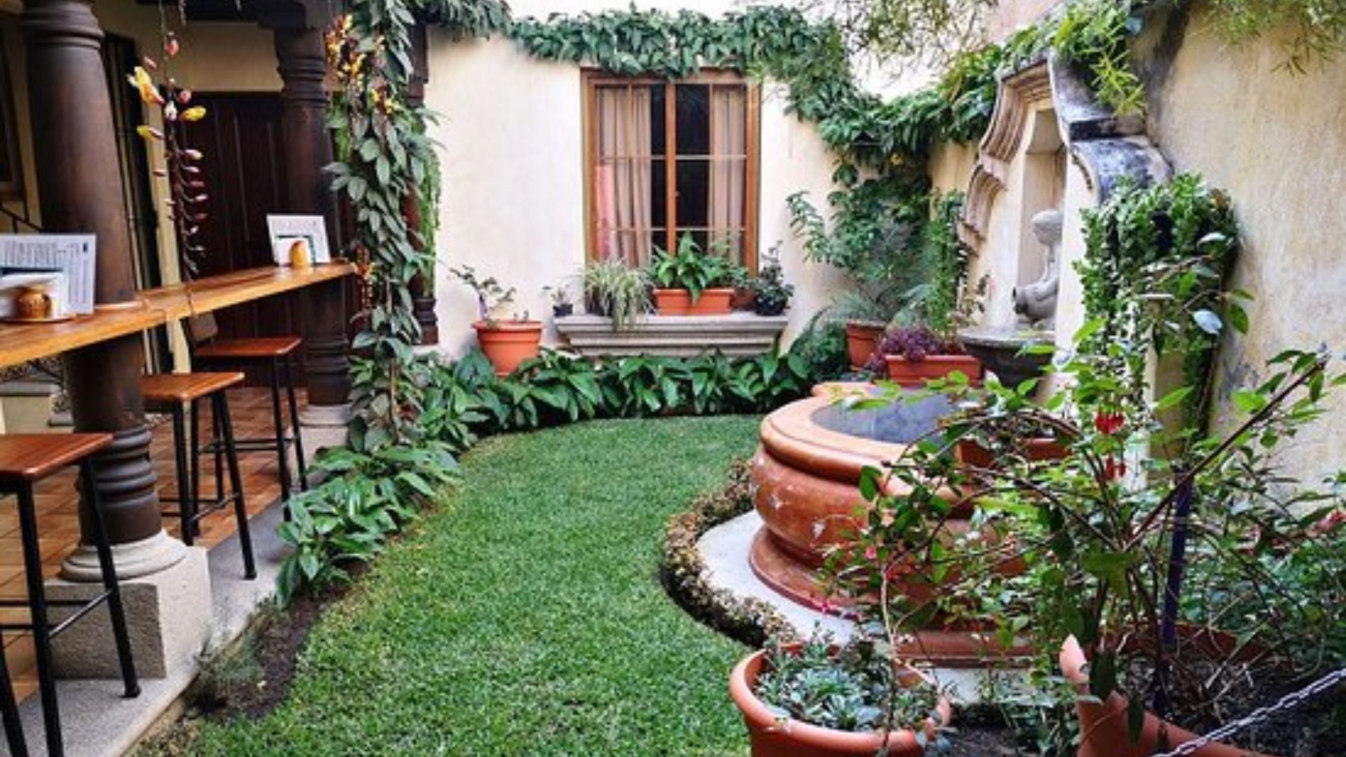 Charming garden patio with lush greenery, a central round fountain, and terracotta pots. A cozy ambiance enhanced by a wooden bar with stools on the left.
