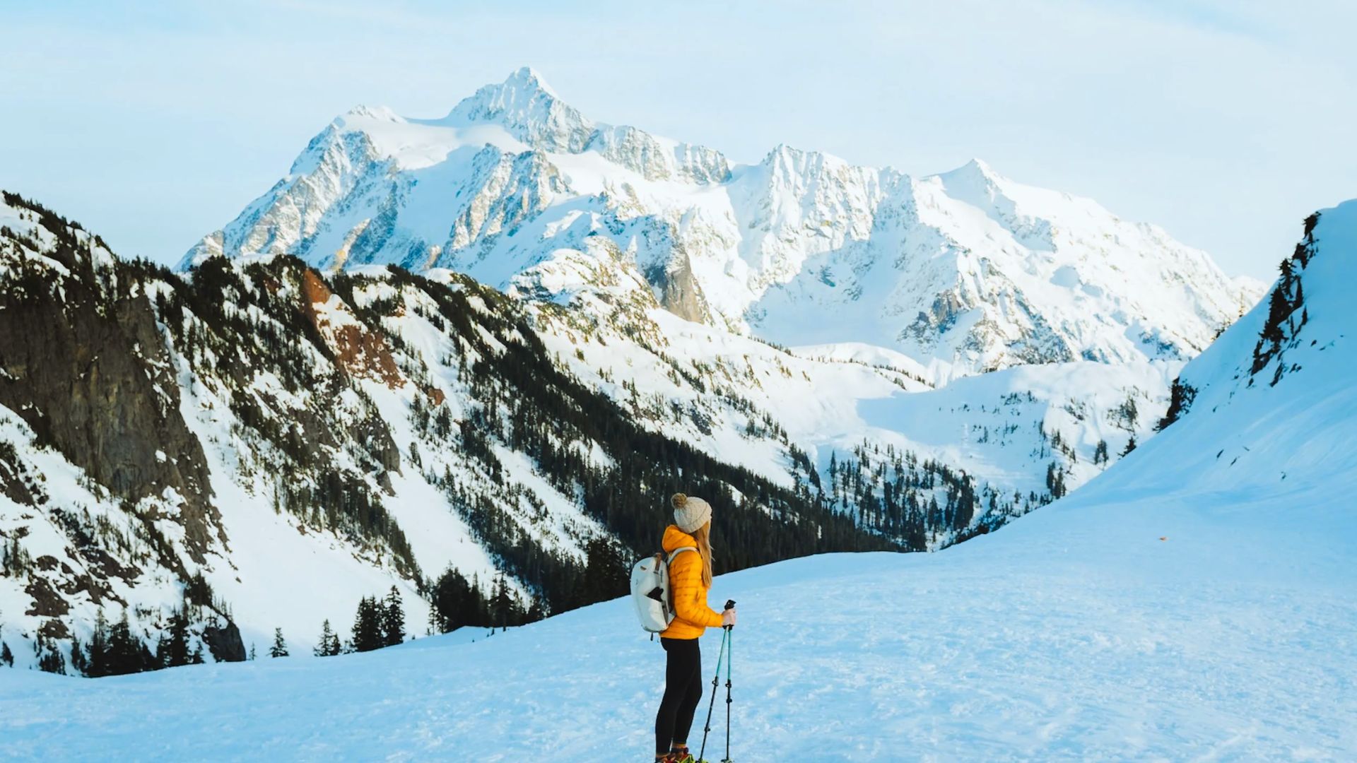 A person in a yellow jacket stands with hiking poles on a snowy mountain, gazing at majestic snow-covered peaks under a clear blue sky. Serenity and adventure.