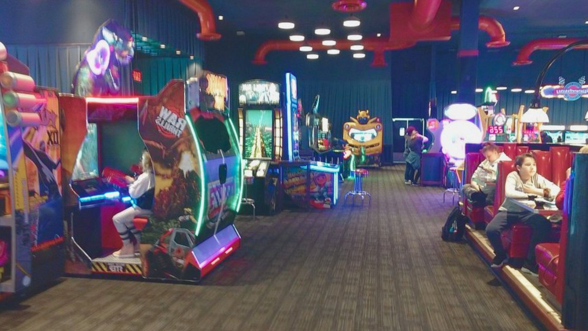 A vibrant arcade with neon-lit game machines and a few people playing or relaxing in booths. The atmosphere is lively and energetic.