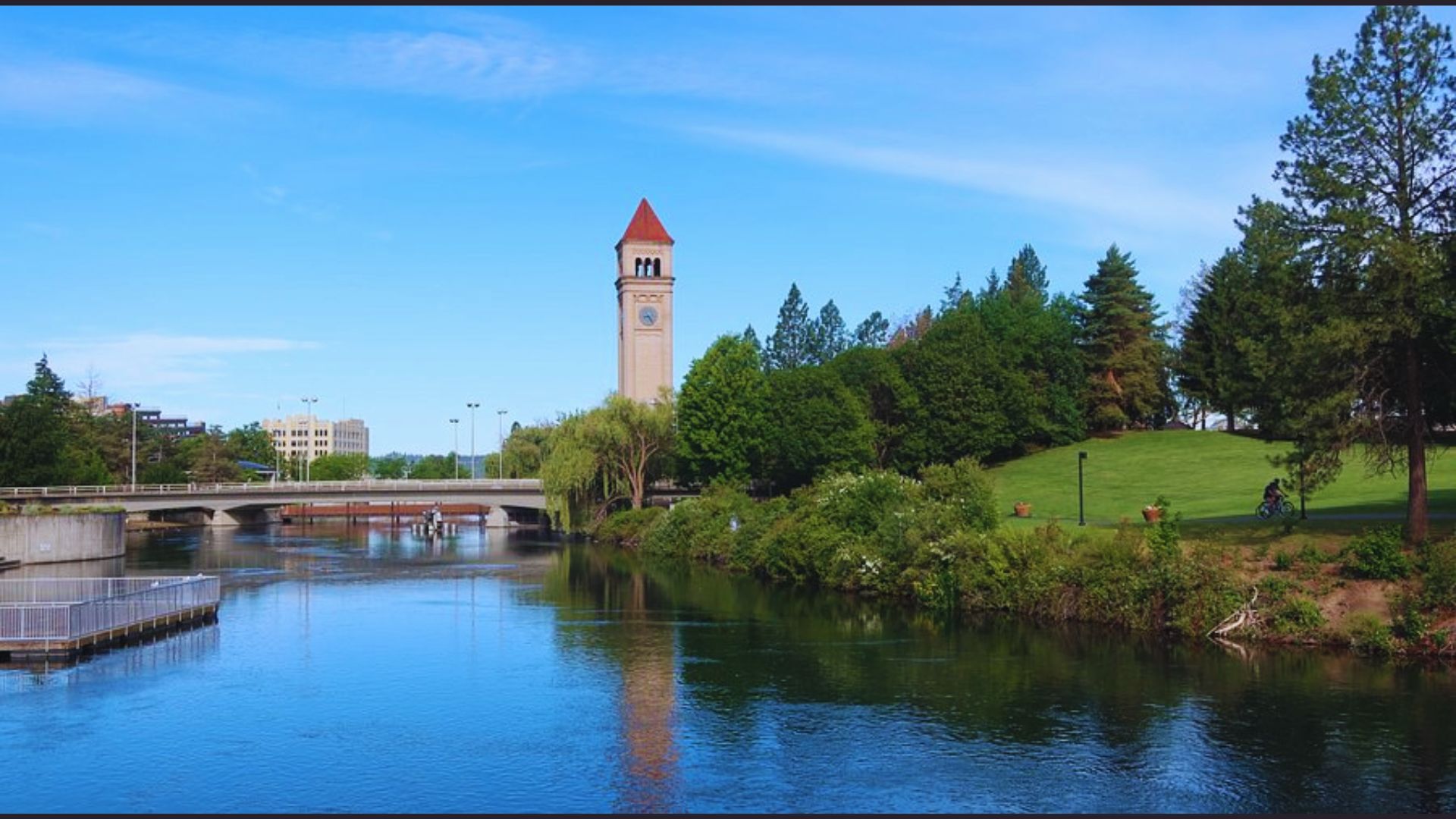 A serene river flows with a clock tower rising in the background against a clear sky.