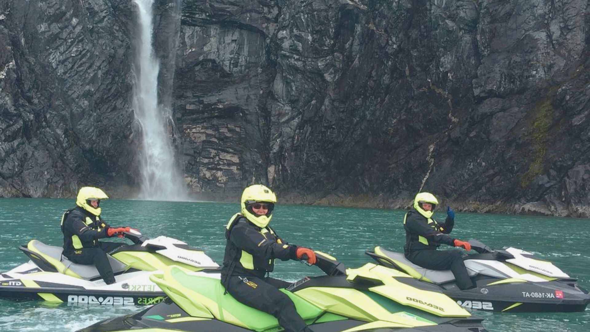 Three people in helmets and gear ride jet skis on a turquoise lake with a tall waterfall cascading down a rocky cliff. The scene is adventurous and energetic.