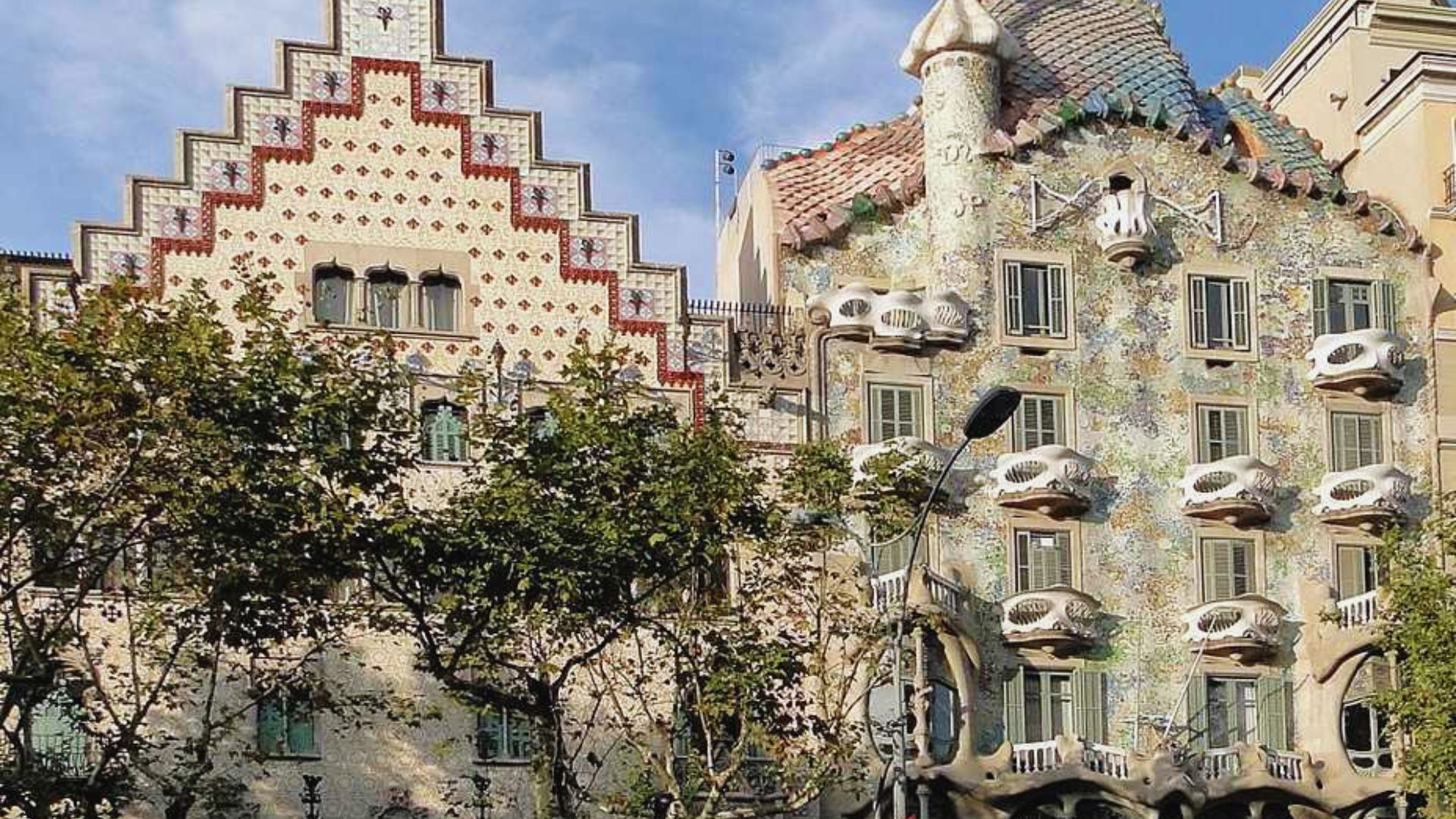 Ornate buildings with colorful, mosaic façades, arched windows, and sculptural balconies. Foreground trees add a natural contrast under a clear sky.