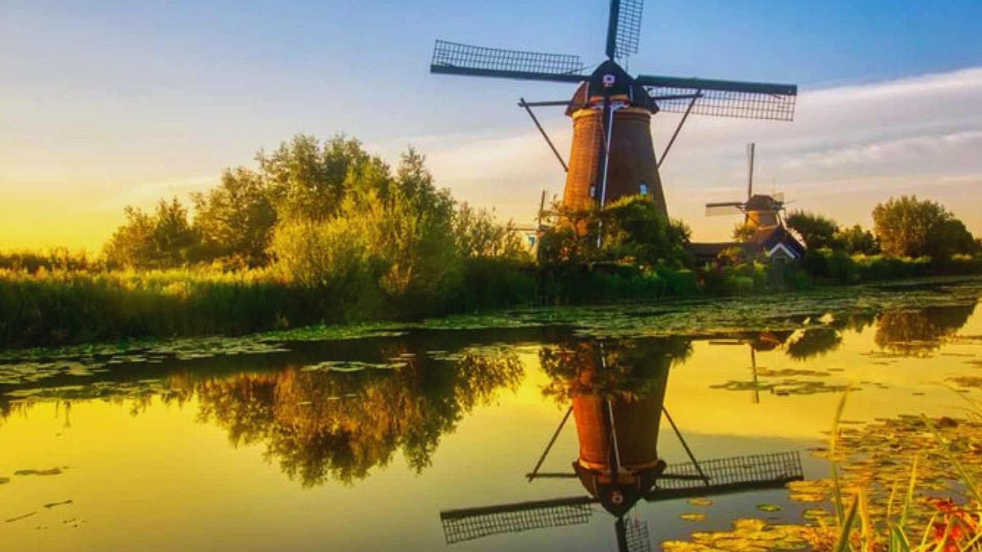 A serene landscape at sunset features two traditional windmills beside a reflective canal, lined with greenery, creating a peaceful, picturesque scene.