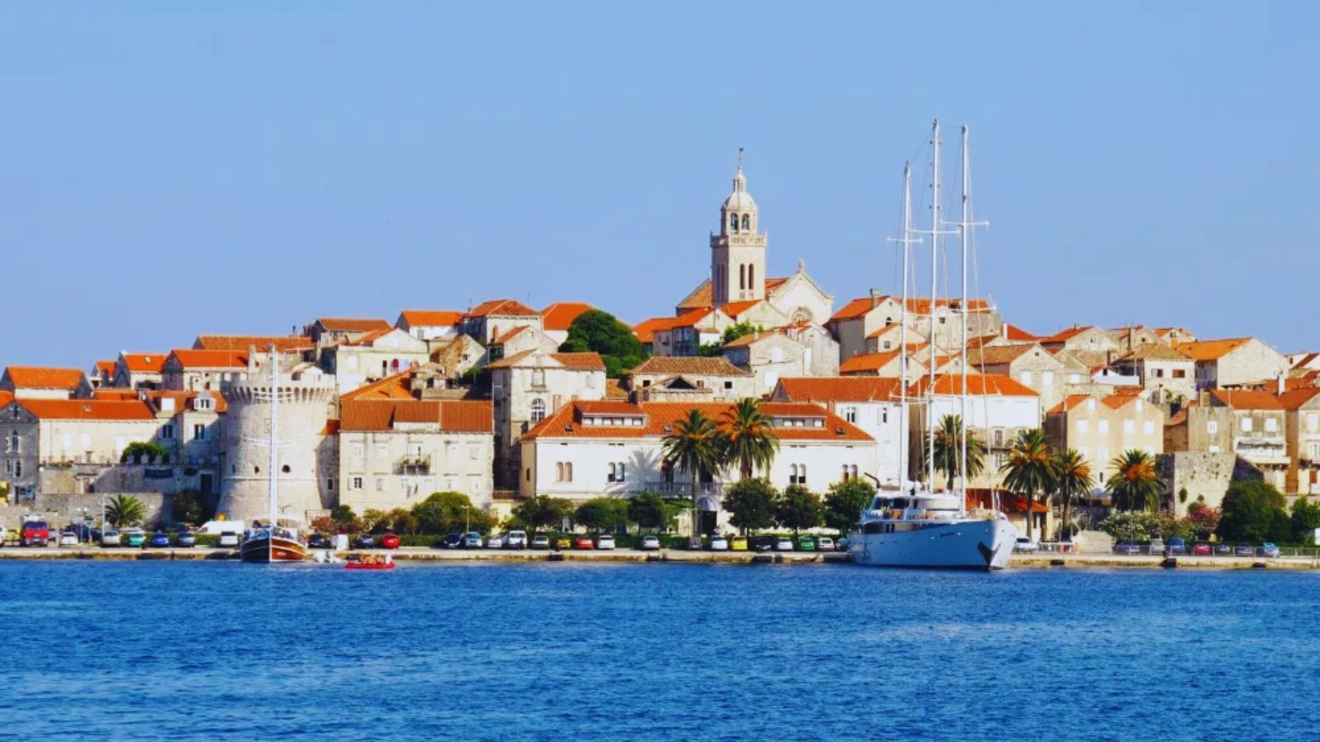 A picturesque coastal town with orange-tiled roofs, a prominent church tower, and calm blue waters. A white sailboat is docked at the shore.