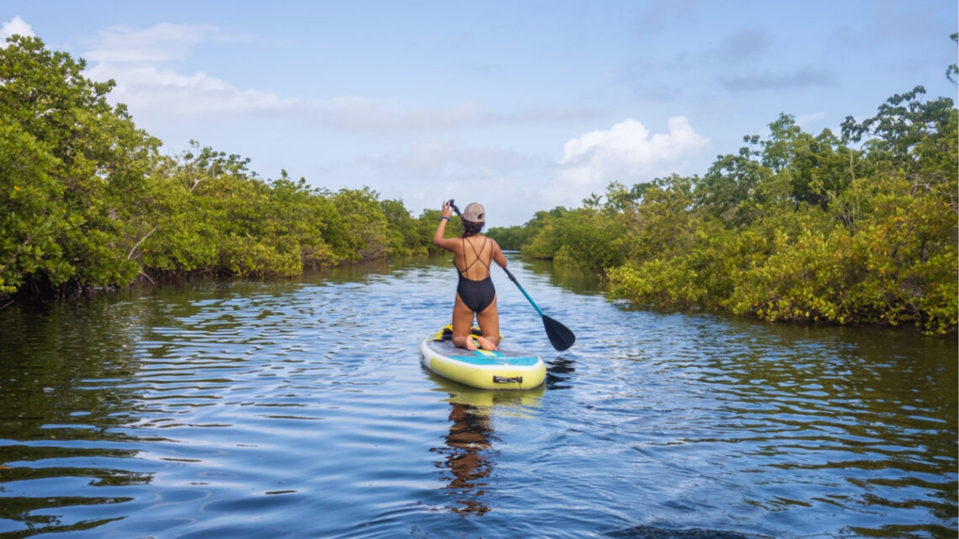 A person in a black swimsuit stand-up paddles through a serene mangrove-lined waterway under a clear blue sky, conveying a sense of adventure and tranquility.