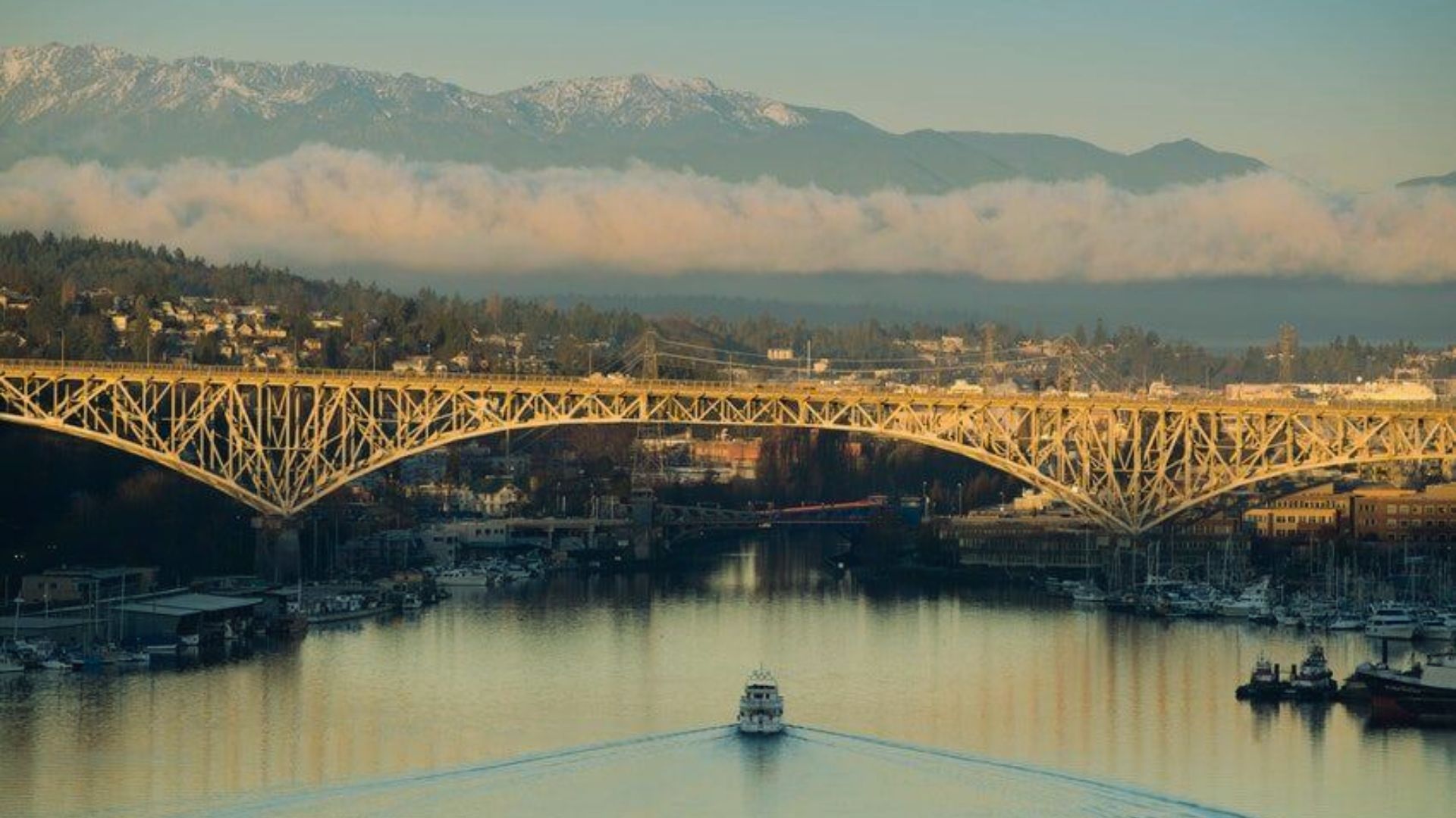 A boat navigates a calm river beneath a large arch bridge at sunset. The background features a town, misty hills, and snow-capped mountains, creating a serene atmosphere.
