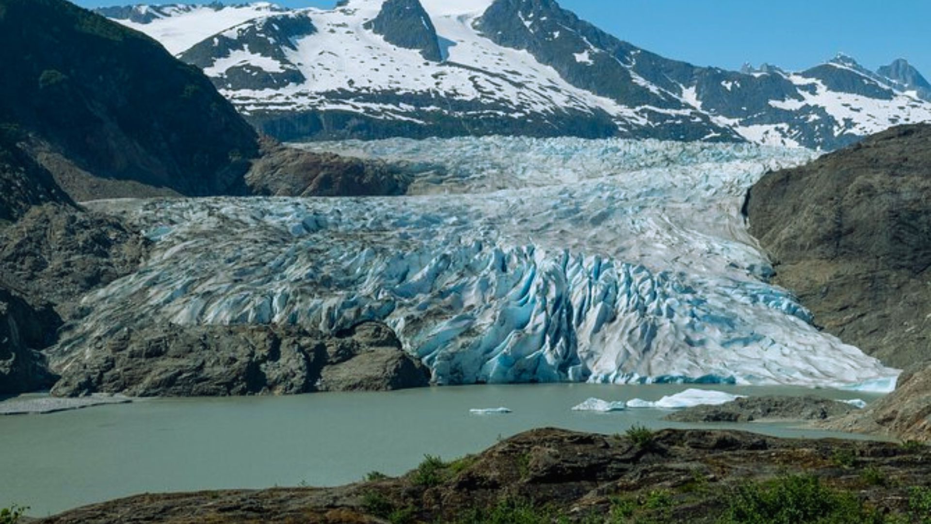 A stunning glacier descends into a serene lake, flanked by rugged mountains. The scene is tranquil, with snow-capped peaks under a clear blue sky.