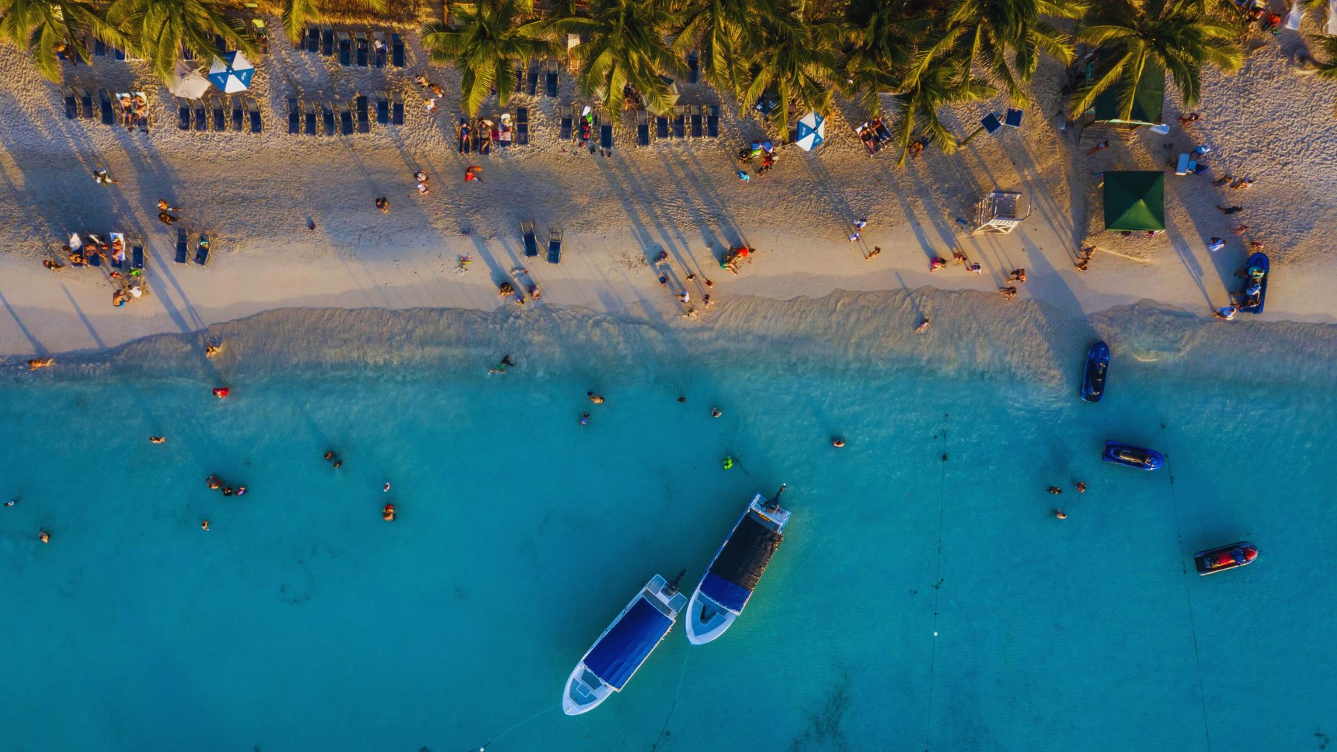 Aerial view of a vibrant beach scene with white sand, turquoise water, boats, lounge chairs, and palm trees. People enjoy swimming and sunbathing.