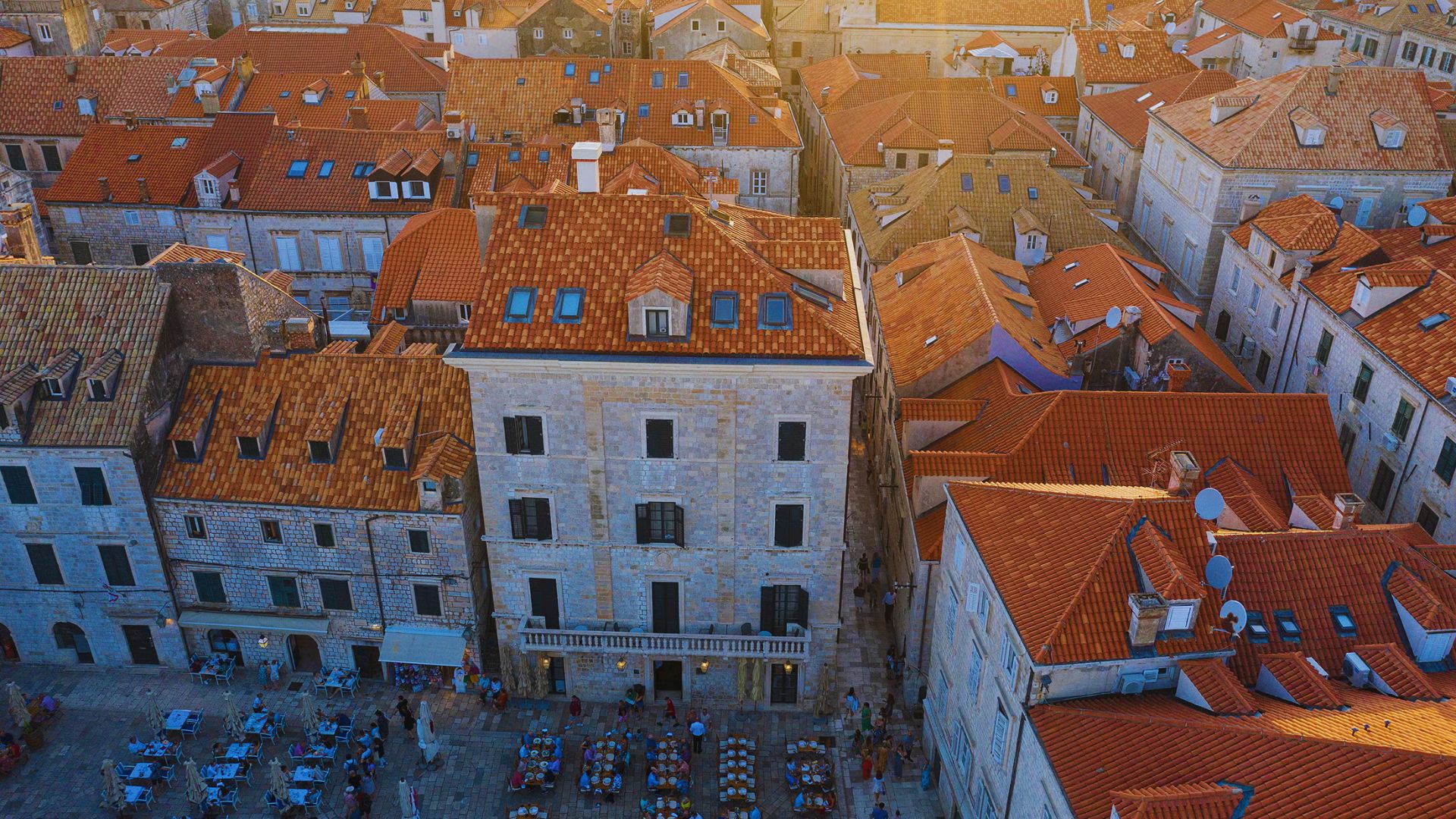 Aerial view of a historic European town with orange terracotta rooftops, narrow streets, and outdoor cafes. Warm sunlight casts a golden glow over the scene.