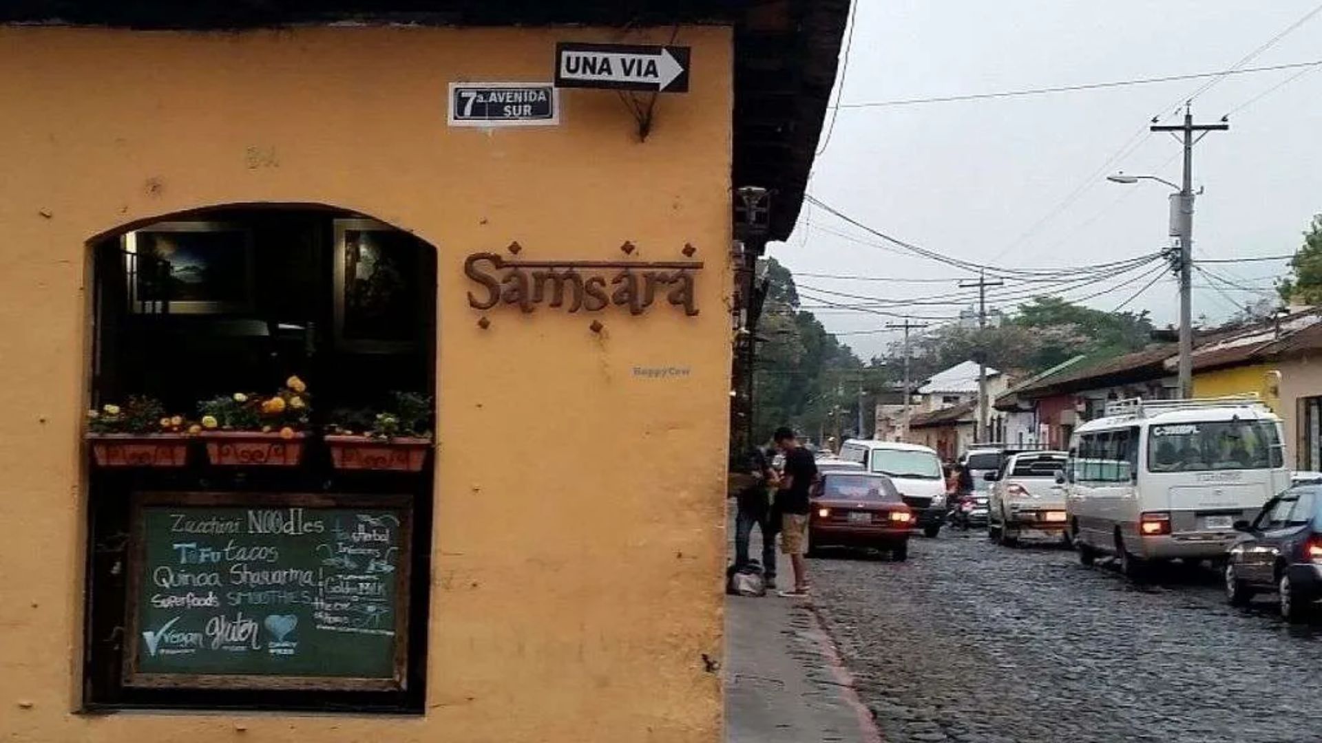 A cobblestone street in Antigua, Guatemala, with a yellow building featuring "Samsara" signage and a chalkboard menu. Traffic and people create a lively atmosphere.