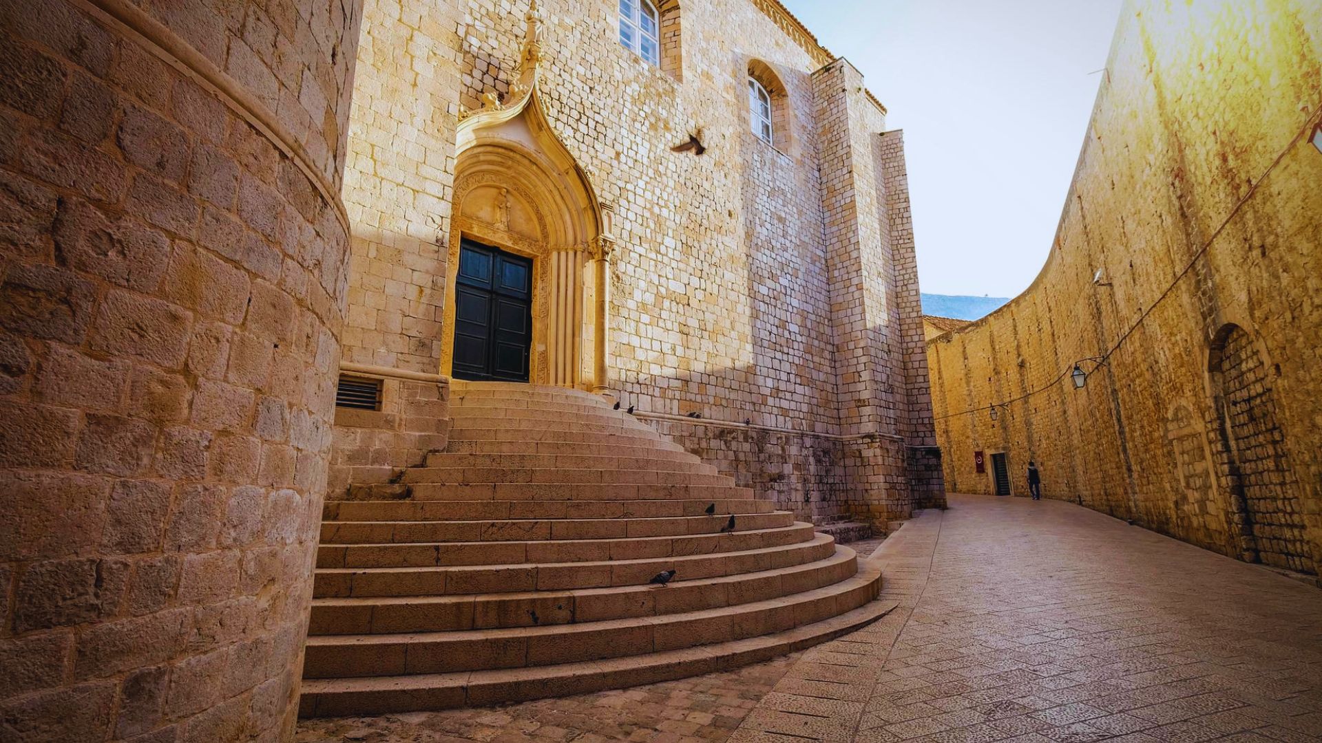 Stone staircase leading to an ornate arched door, flanked by tall historic walls. The warm tones and narrow street evoke a sense of ancient tranquility.